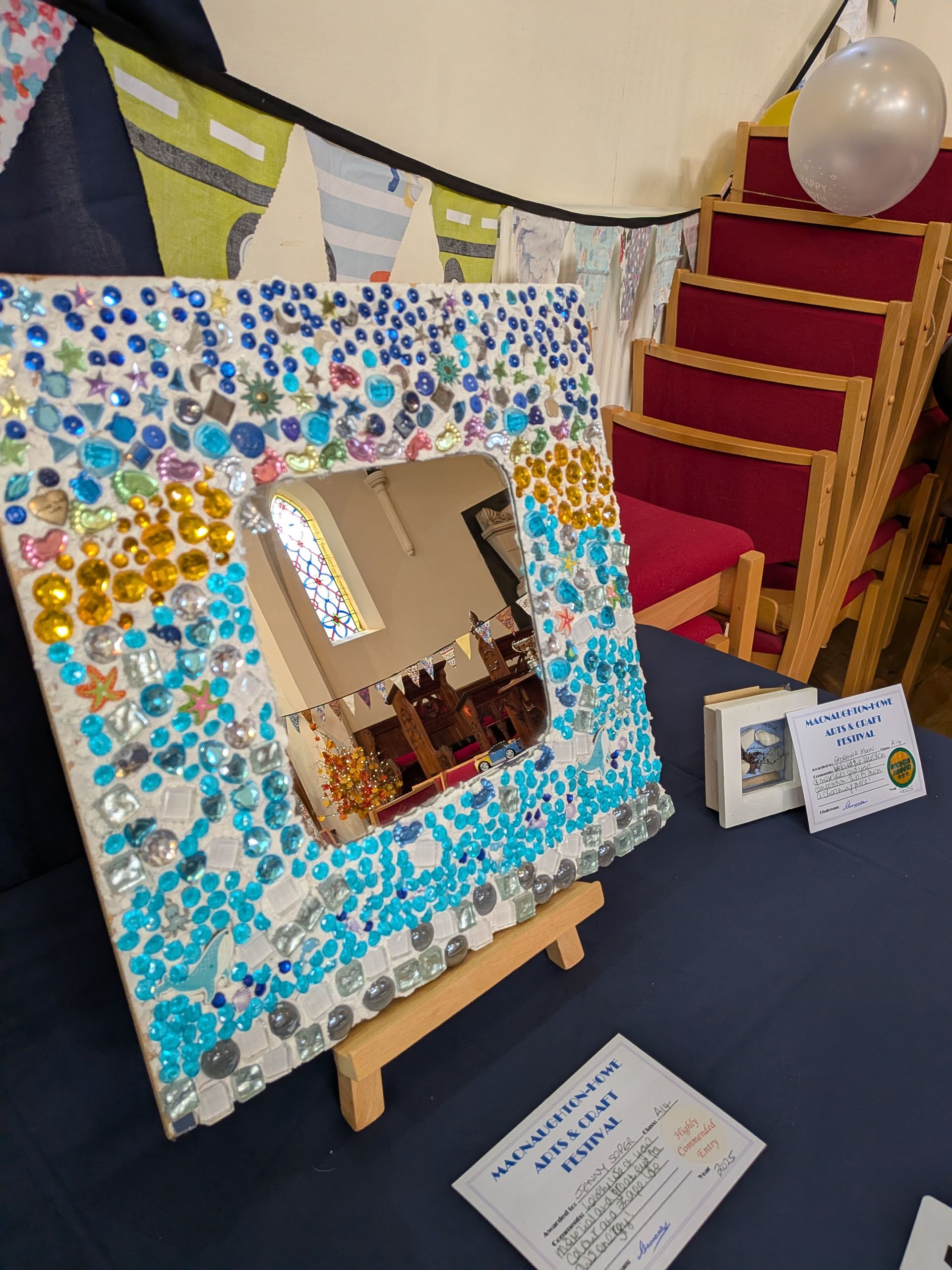 Mosaic-framed mirror on a small easel, reflecting a room. Displayed on a blue table with other objects.