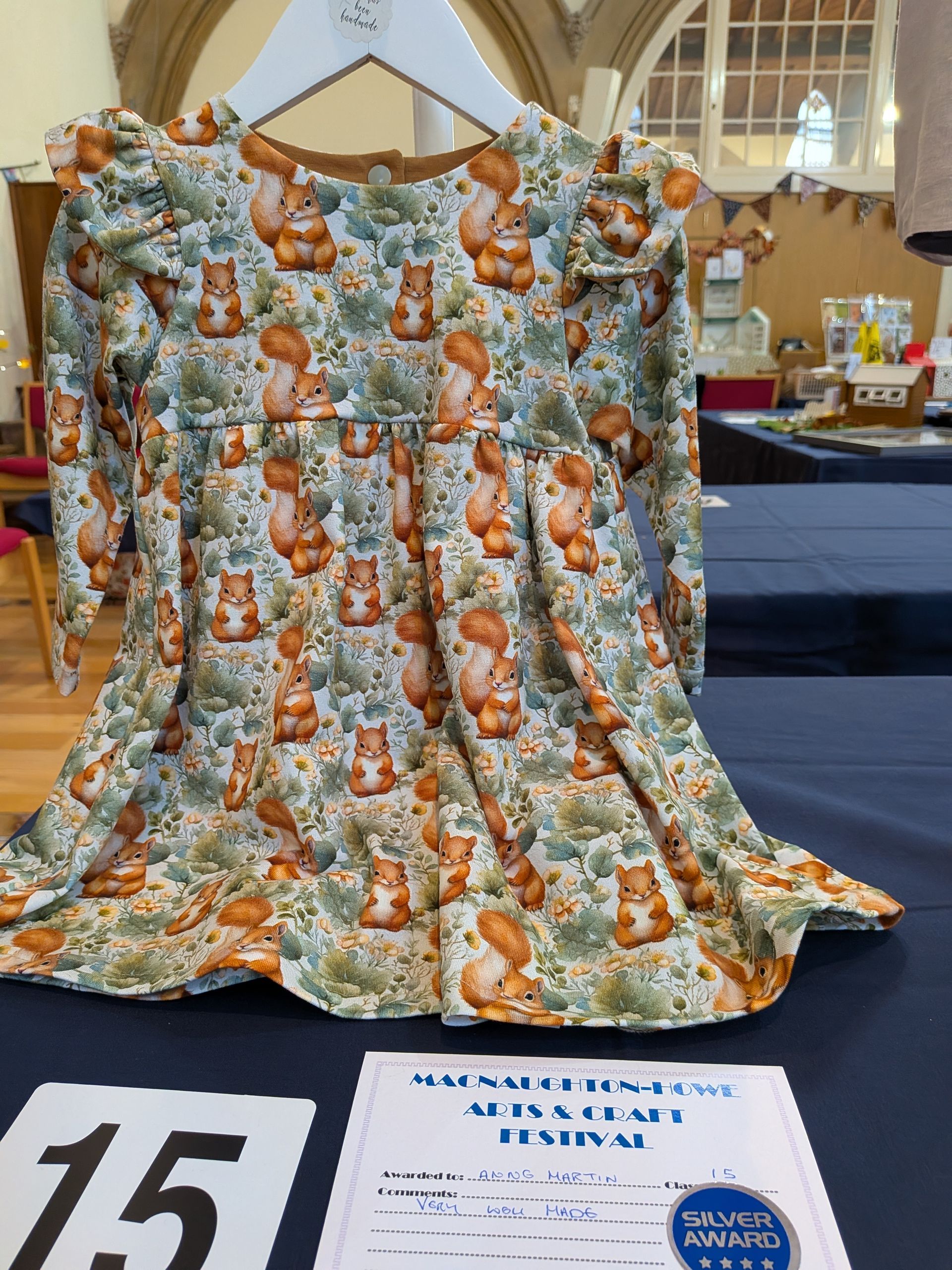 Squirrel-patterned dress on a hanger, displayed on a table. The dress has long sleeves and ruffled shoulders.