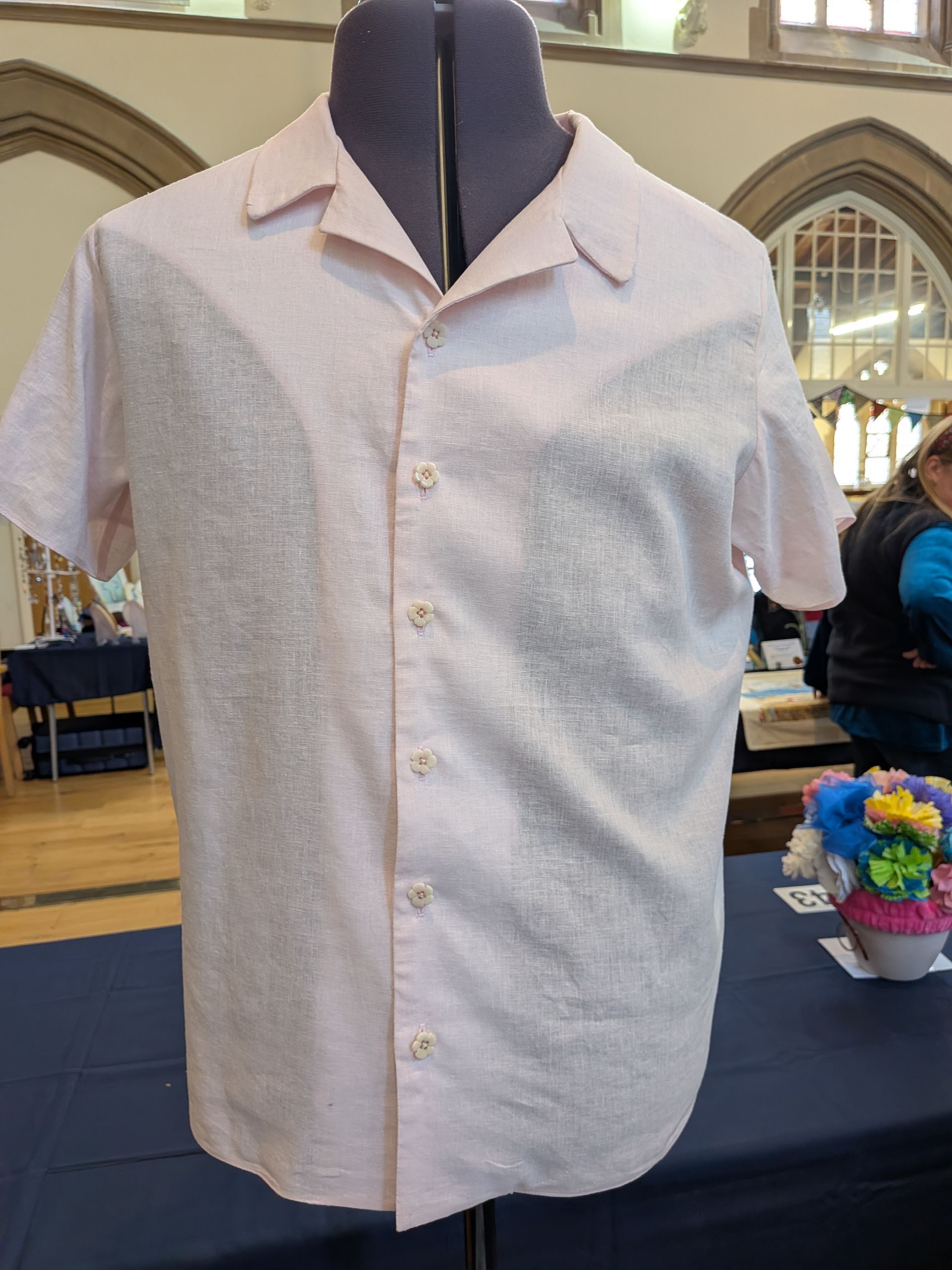 Pink collared shirt on a mannequin. It has short sleeves and is buttoned. The background is indoors with tables.