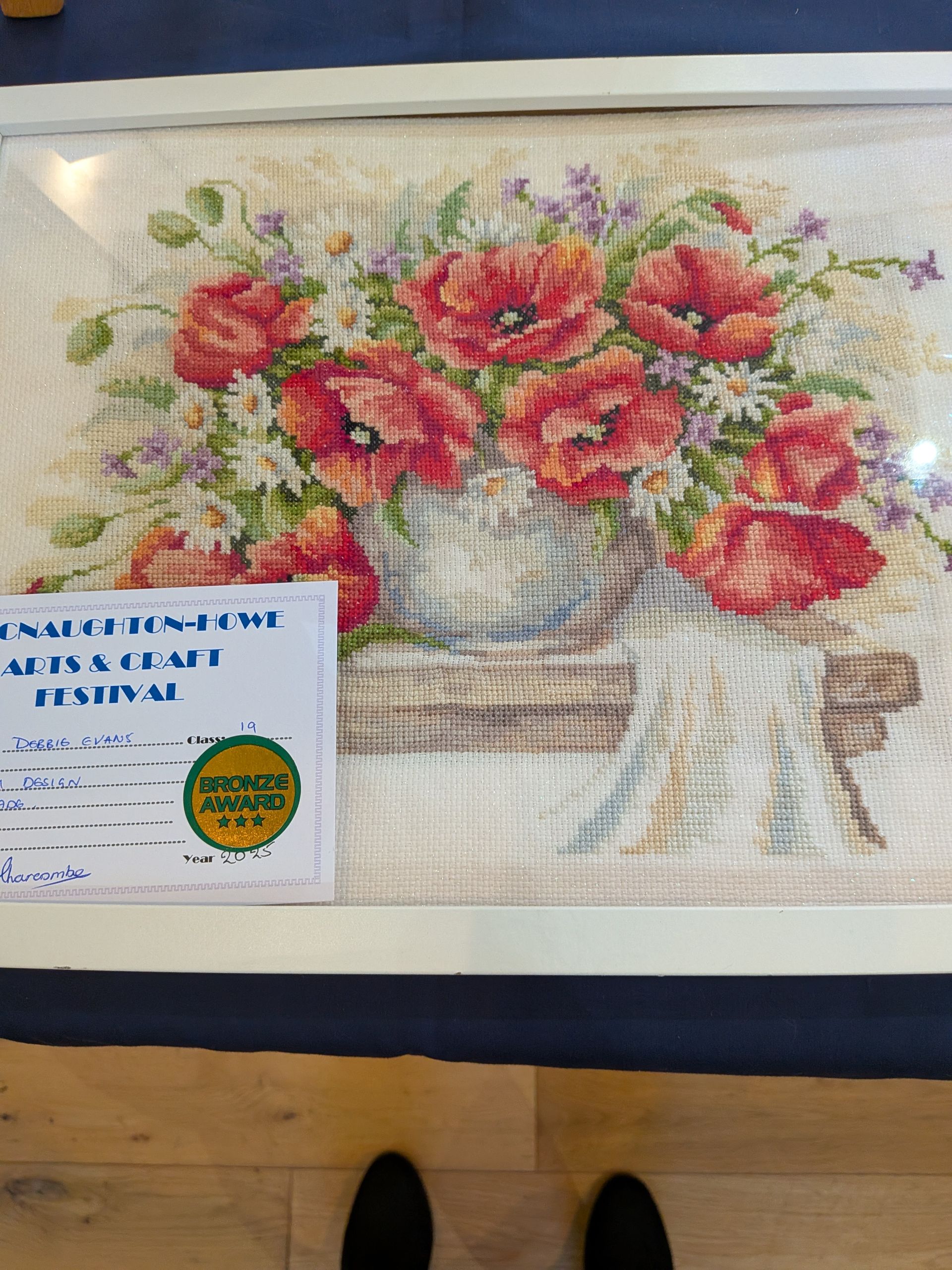 Cross-stitch floral display featuring red poppies in a vase, on a table. White framed, with festival signage.