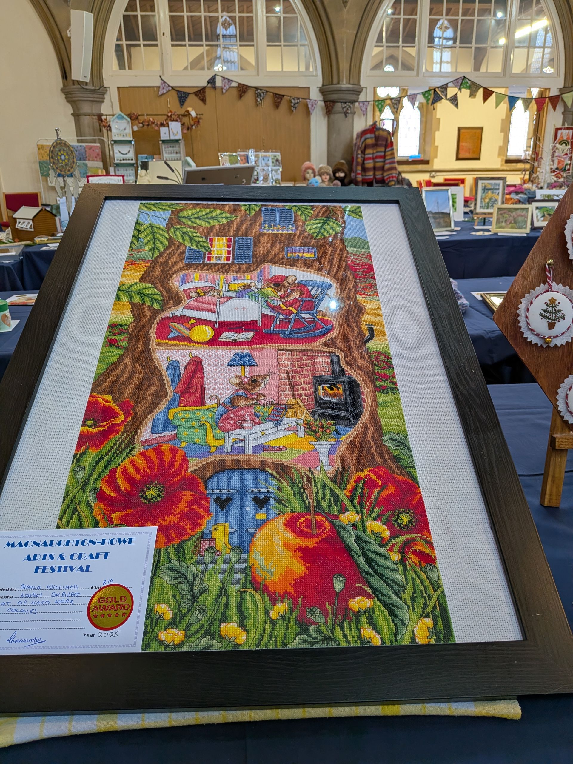 Cross-stitch art of a tree house framed on display at a craft fair. Vivid colors, poppy flowers, and detailed interior scenes.