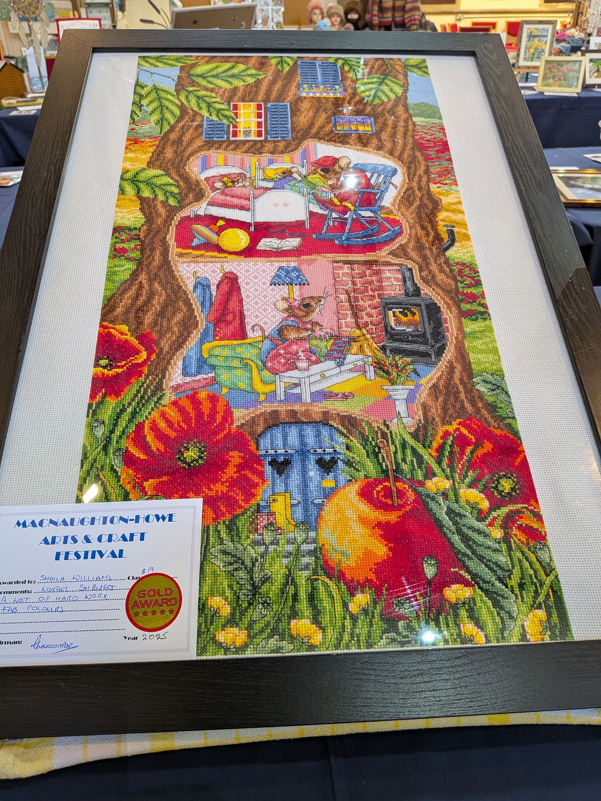 Framed cross-stitch art: tree with open interior revealing whimsical scenes, vibrant colors, display at event.