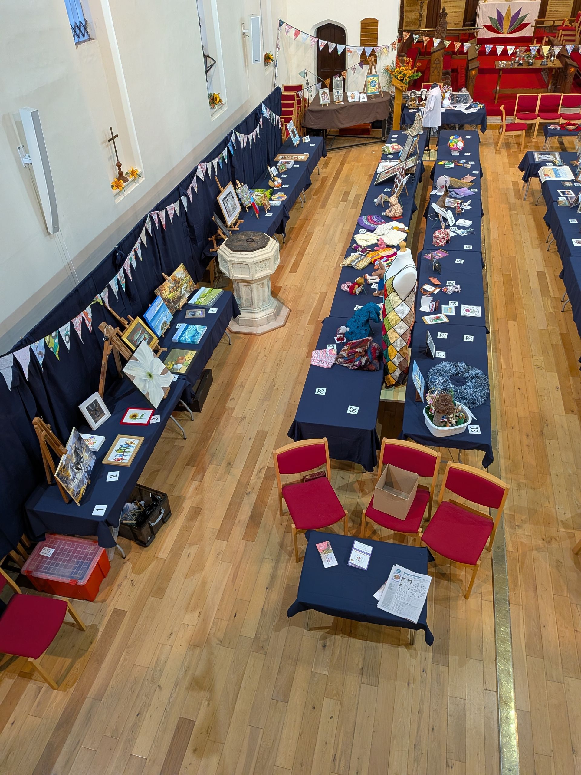Tables with various crafts and art are set up inside a building. Red chairs are placed around some tables.