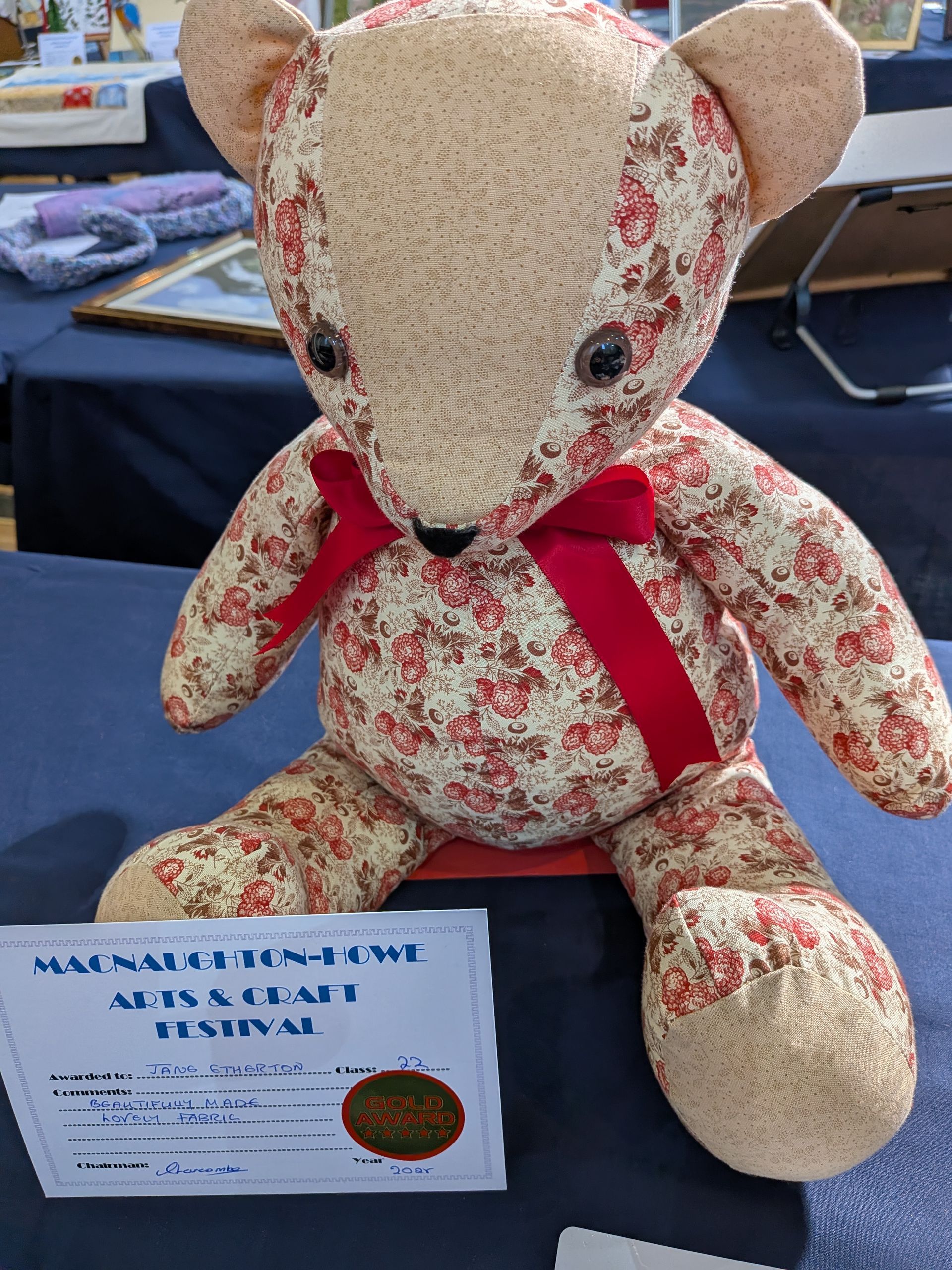Teddy bear with floral fabric, red bow, sitting on a table with a festival sign.