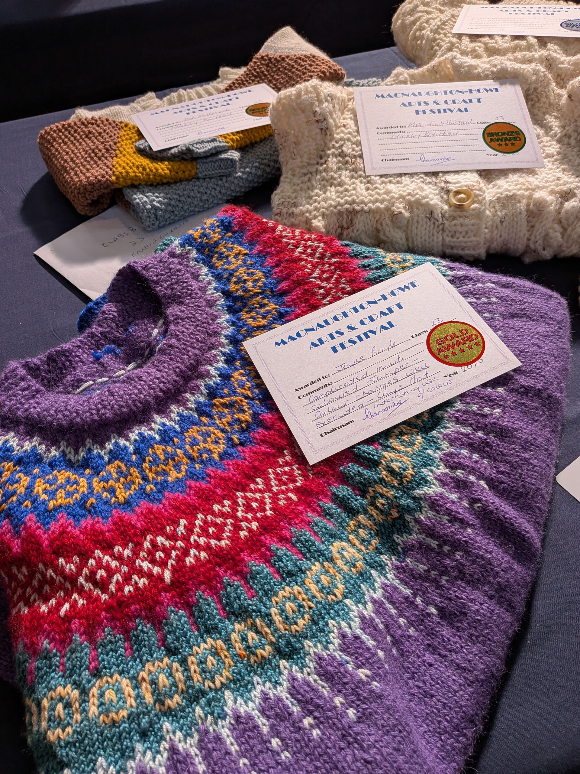 Close-up of colorful knitted sweaters stacked on a dark surface. Each has a tag.