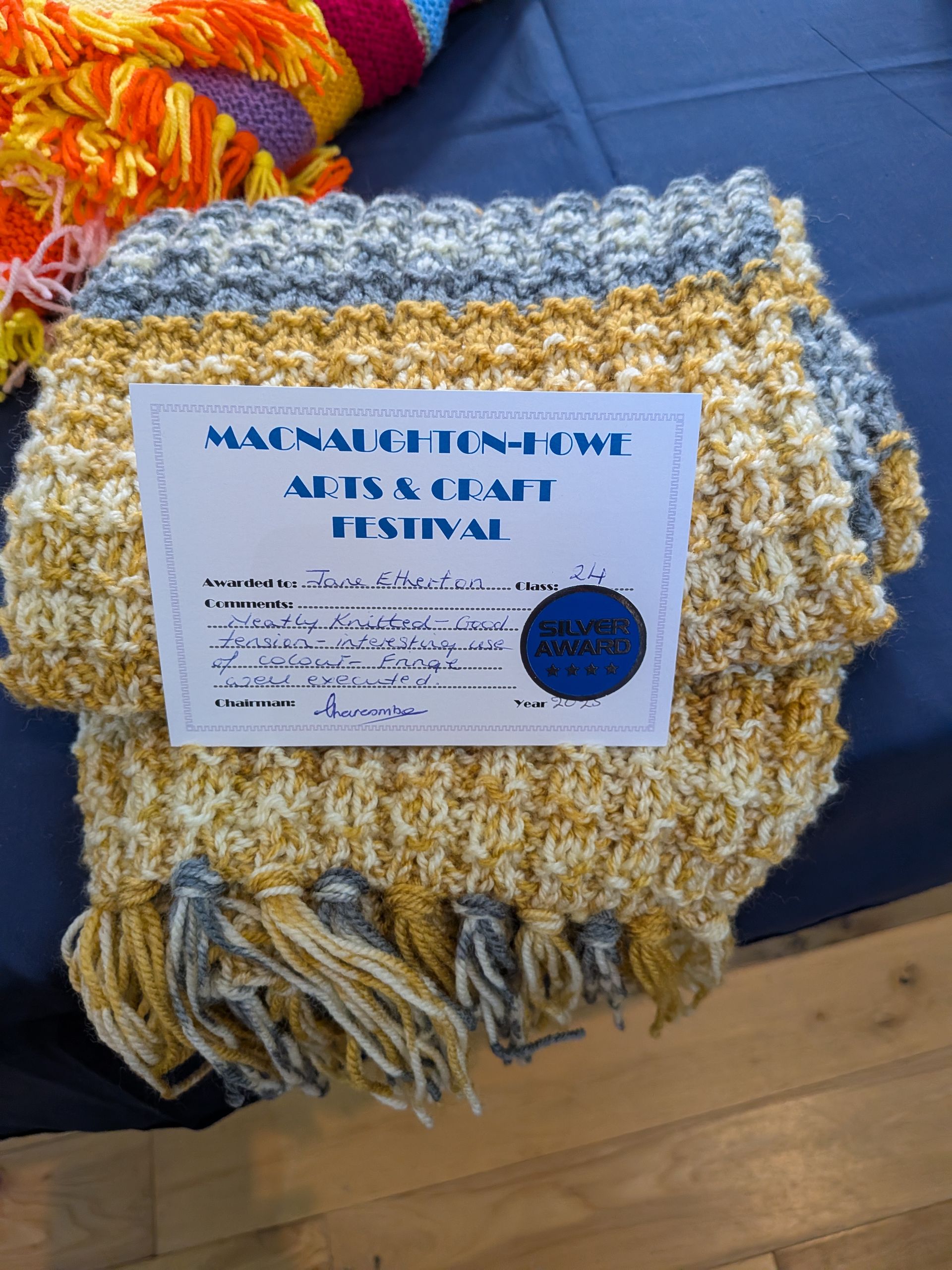 Crocheted scarf in yellow, gray, and white with attached festival information.