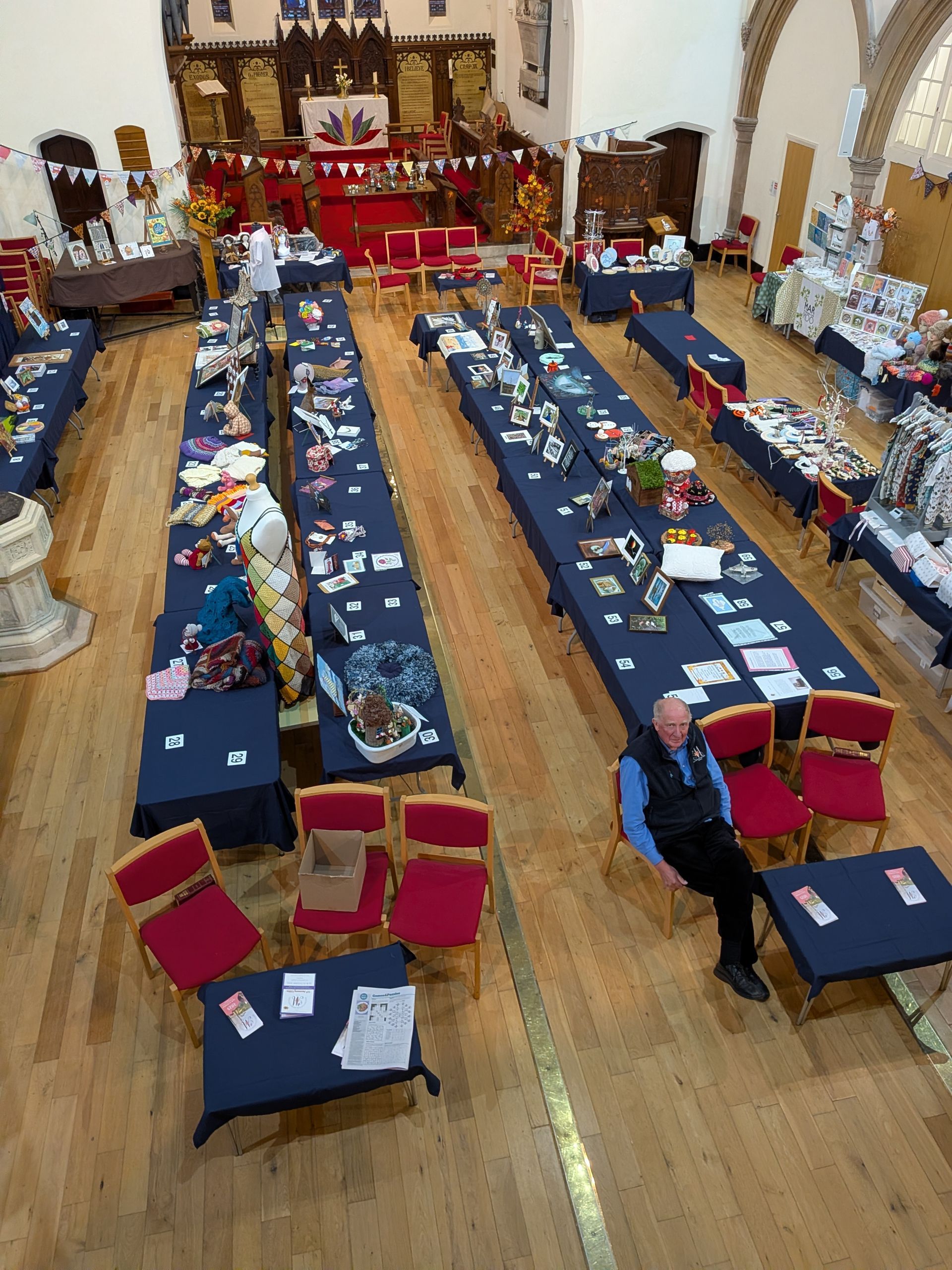 Long tables with crafts at a church fair.  Man in chair at one table.