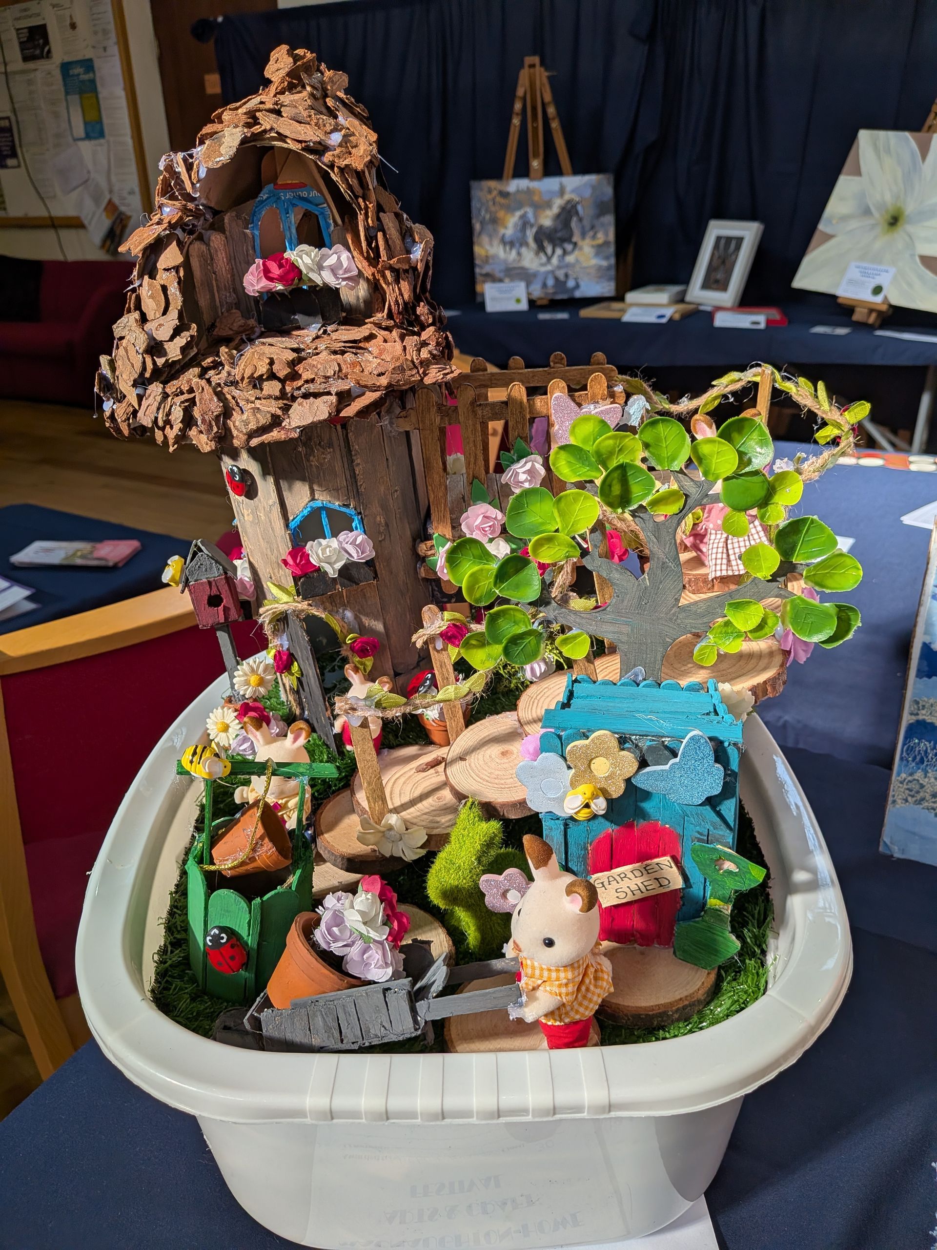 Fairy garden diorama in a white container, with house, trees, figures, flowers, and other whimsical details.