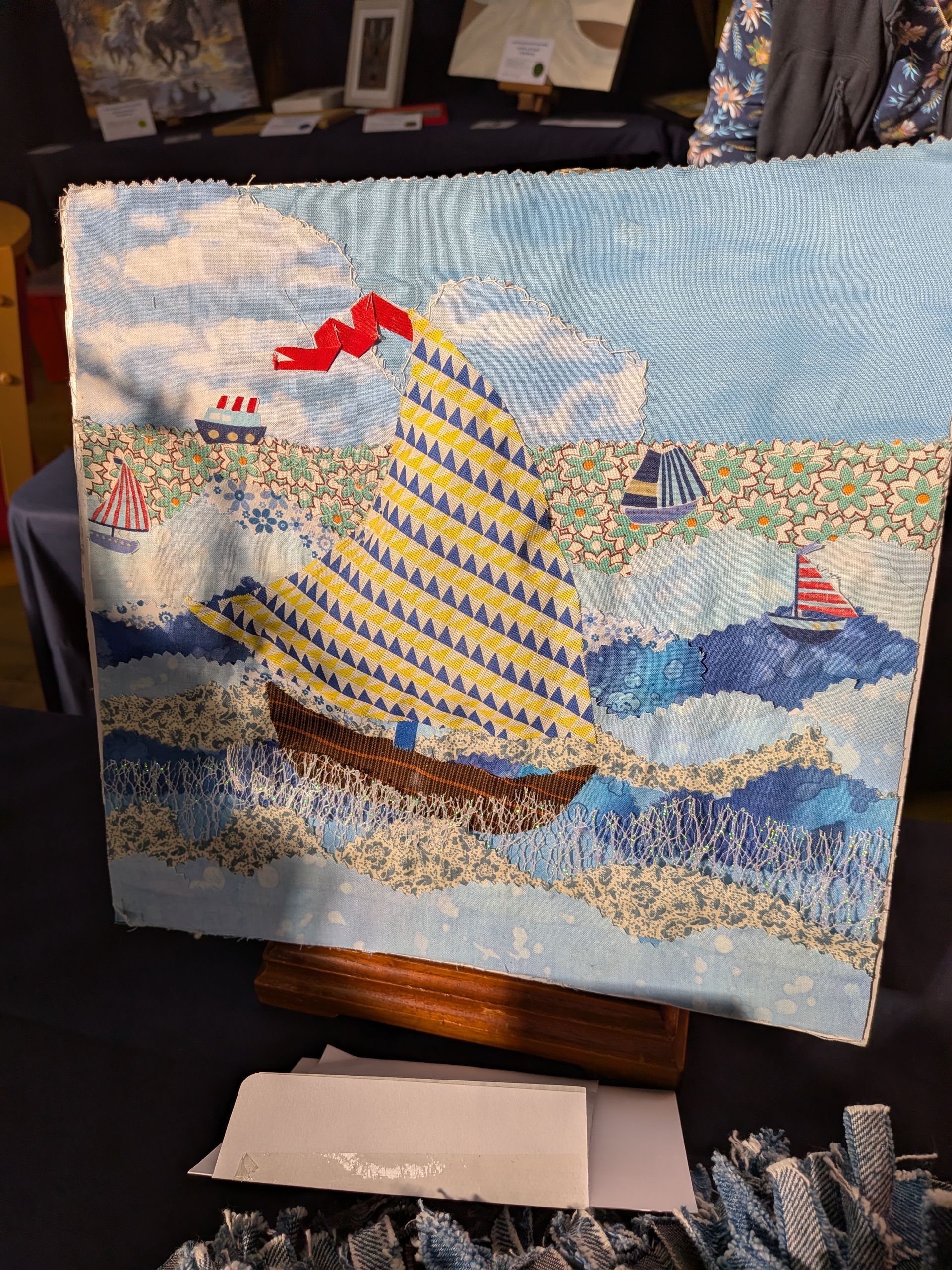 Needlepoint of a sailboat with yellow sail and red pennant, sailing on blue waves, two other ships visible.