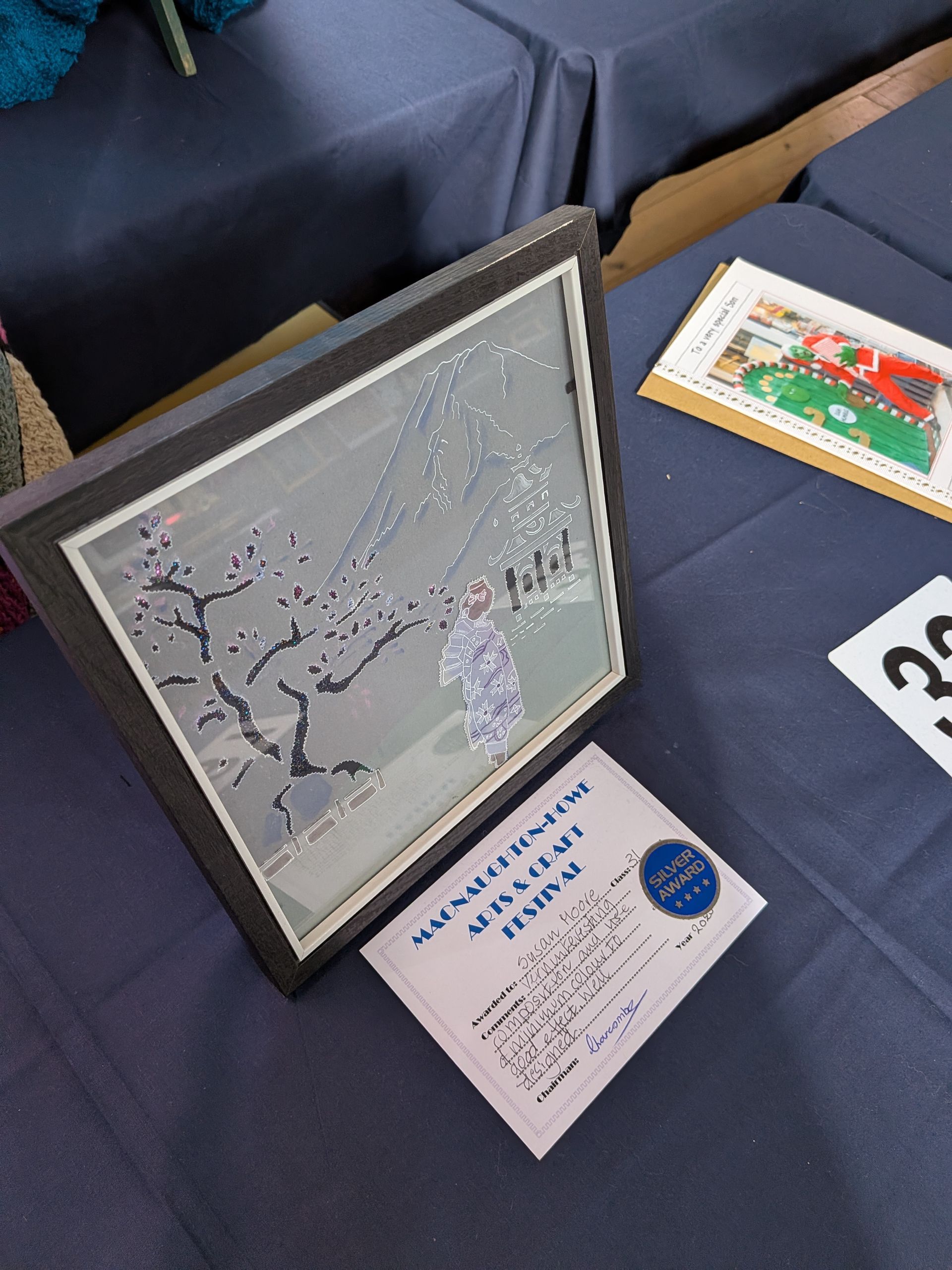 Framed artwork featuring a figure under blossoming branches, on a table at an art festival.