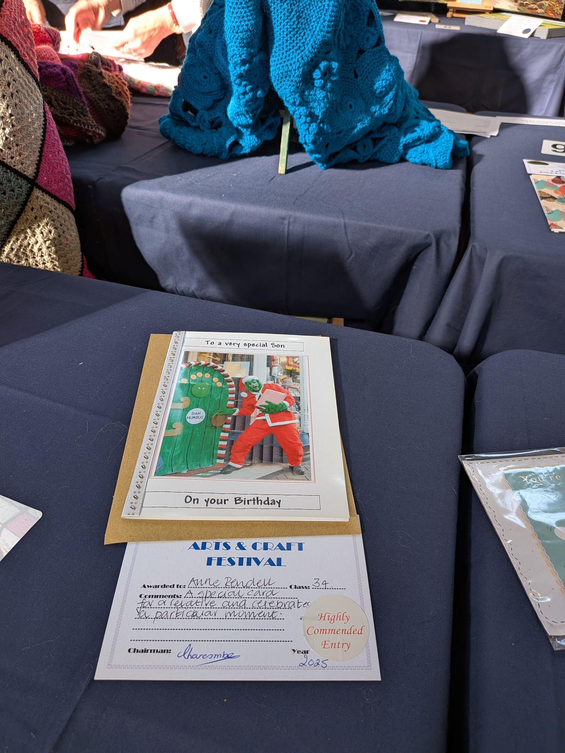 A craft festival display: pattern book showing a green and red costumed duo, blue knitted items, and price lists.