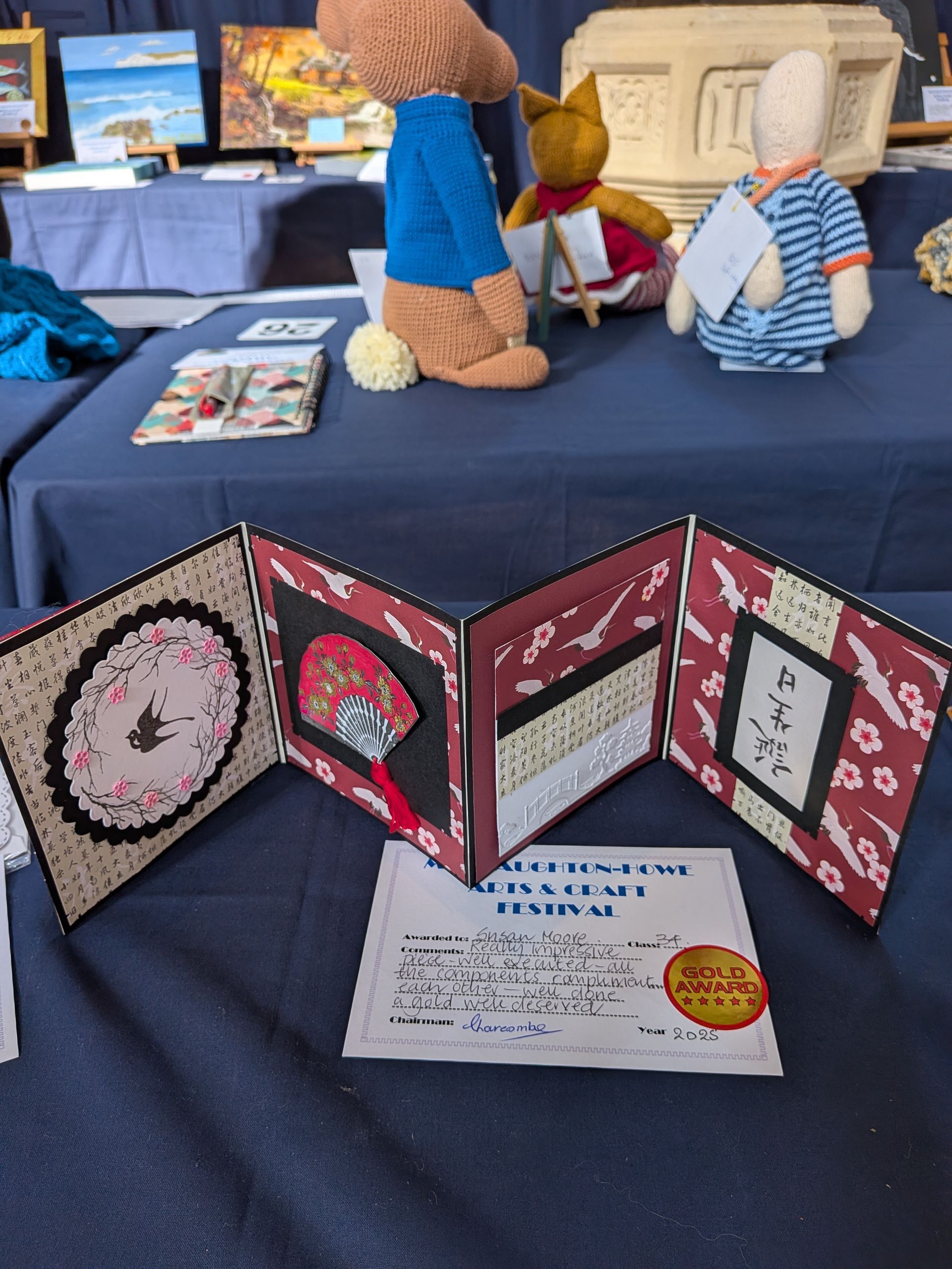 Accordion-style artwork on a table, with stuffed animal figures in the background. A certificate lies in front.