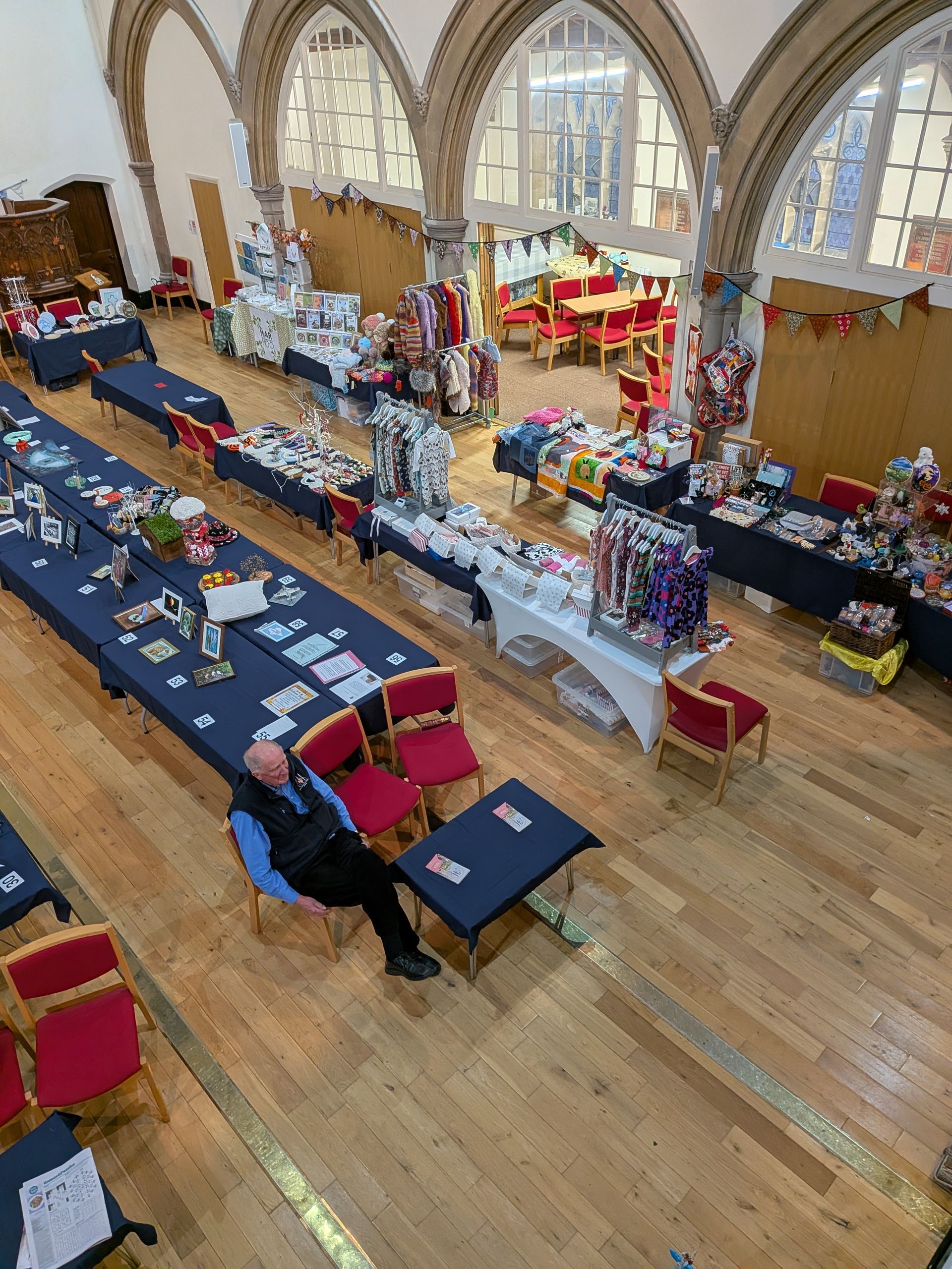 A man sits at a table in a hall with several vendor tables. The hall has high arched windows.