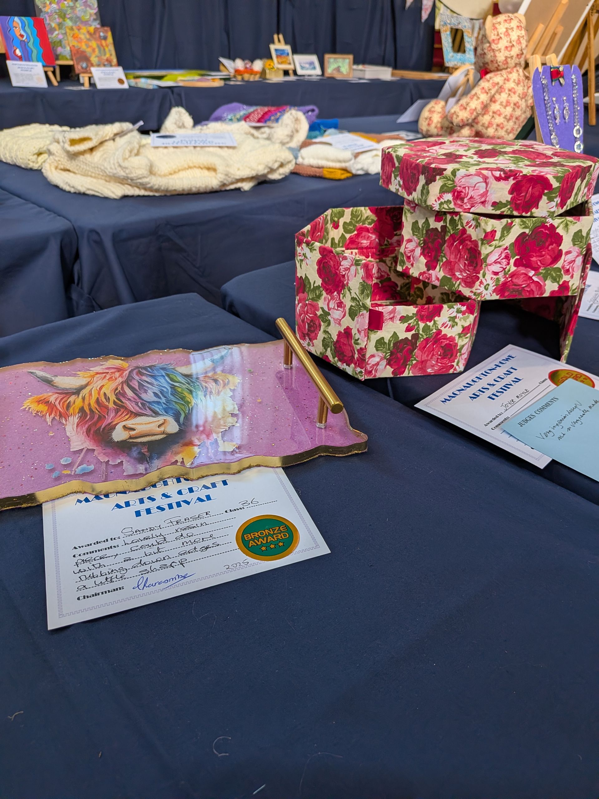 A craft fair table with artwork, a floral box, and a painting of a Highland cow.