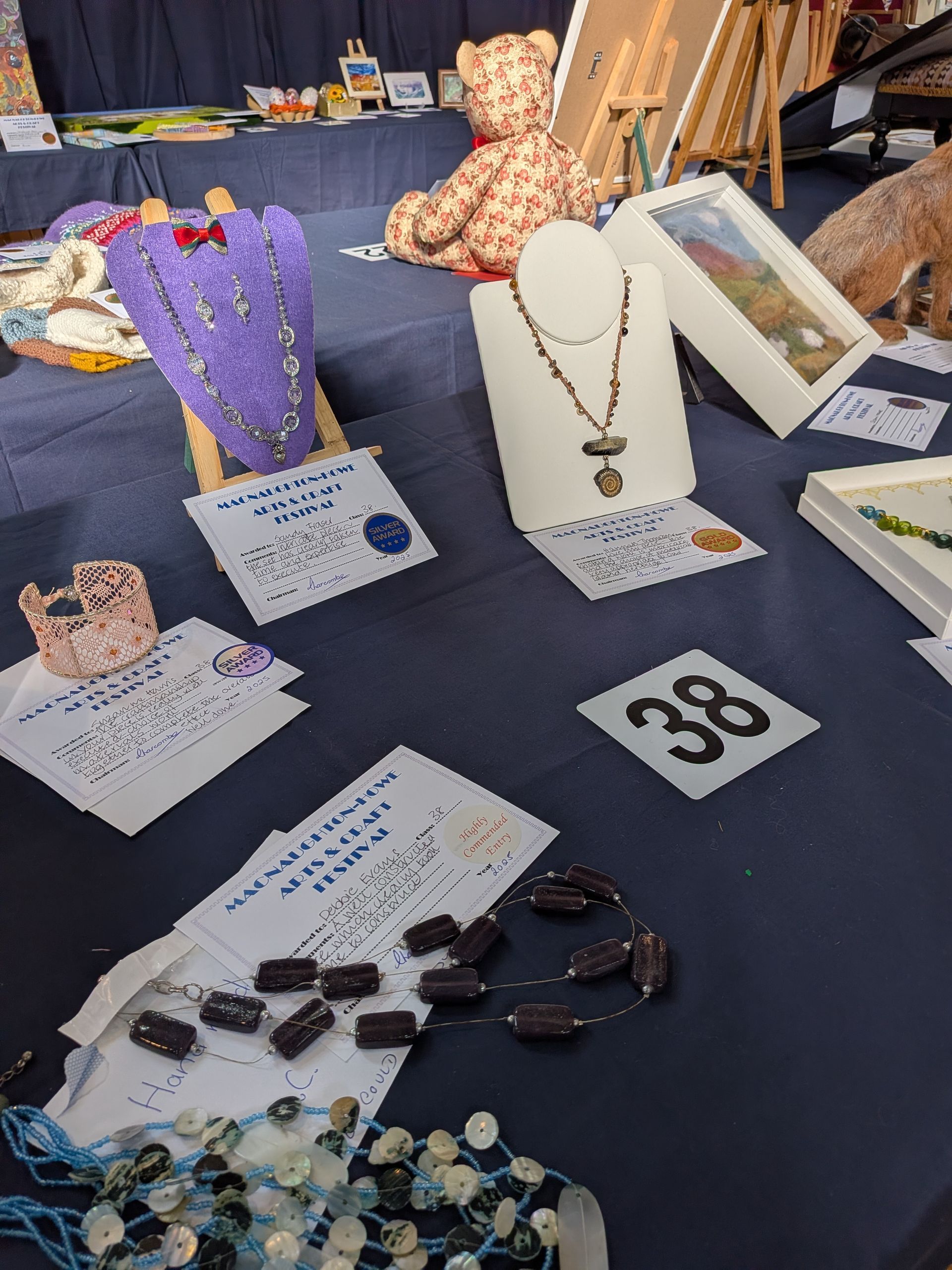 A craft fair table displays jewelry, art, and a stuffed bear on a blue tablecloth.
