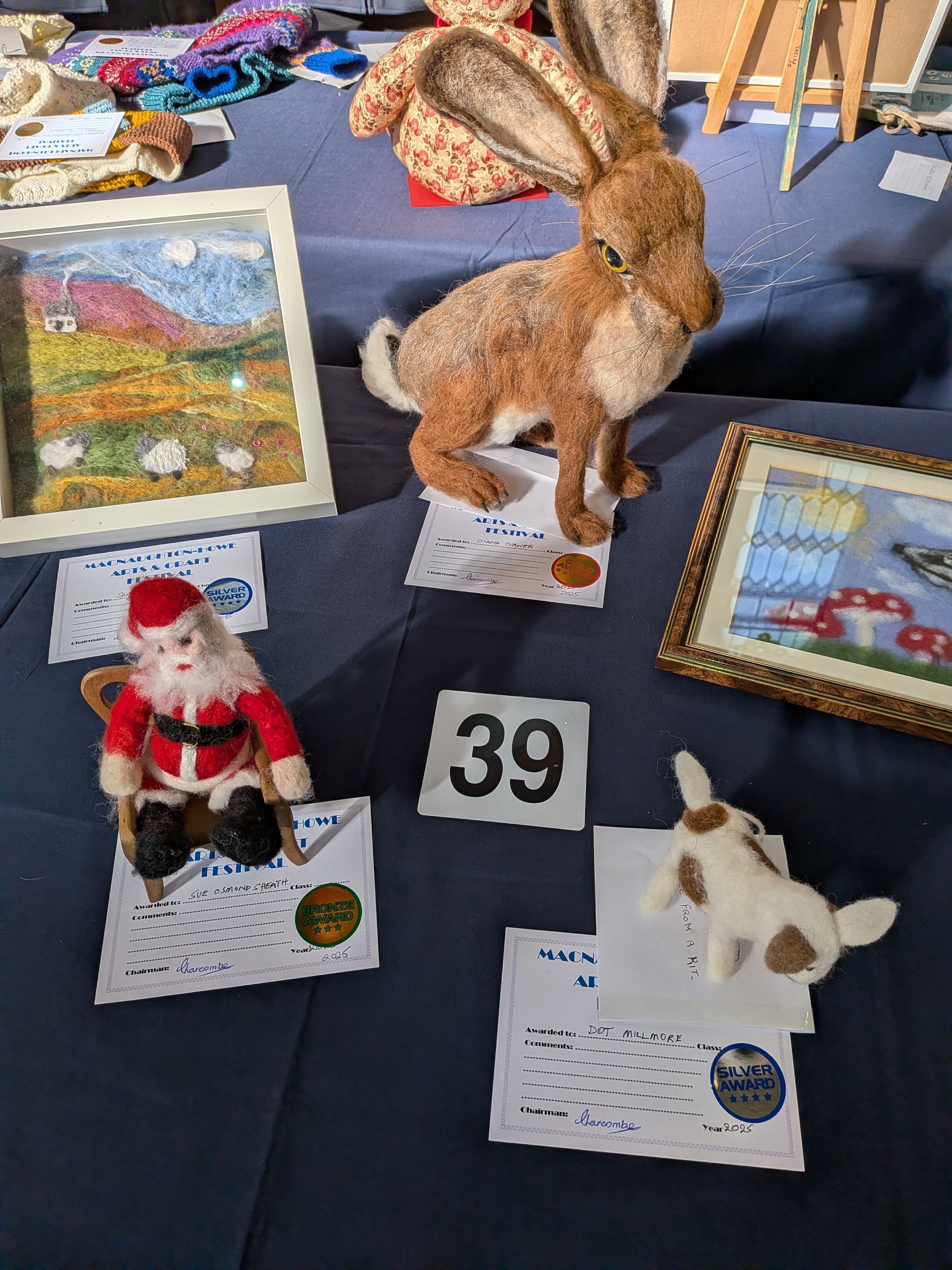 Craft fair display: a Santa figure, a rabbit, and two framed needlepoint pieces, all on a blue tablecloth.