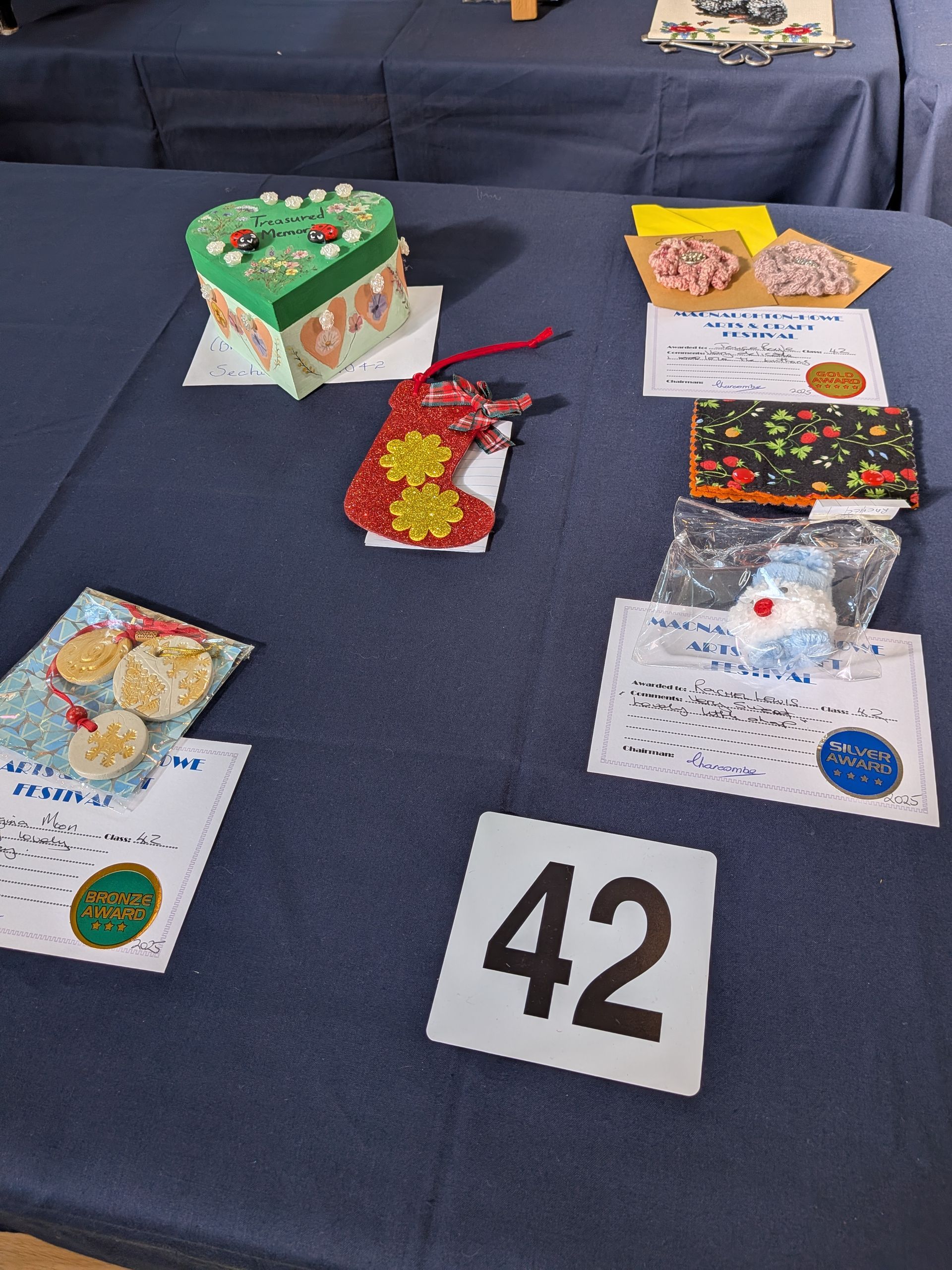 Items on a blue tablecloth, including a heart-shaped cake, cookies, and other crafts with labels. Number 42 in foreground.