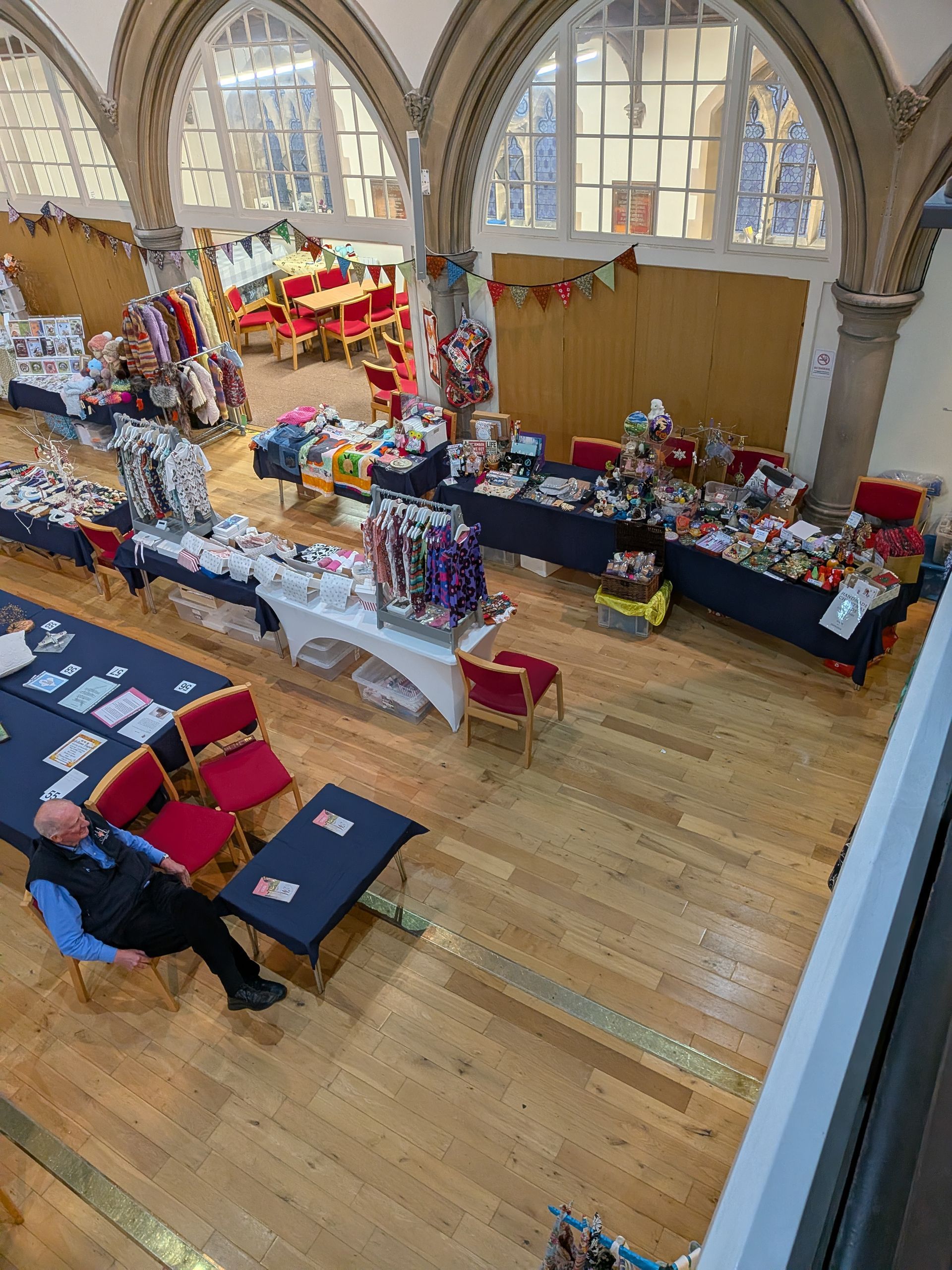 An overhead view of a hall with tables displaying merchandise. A man sits at a table. Wooden floor, arched windows.