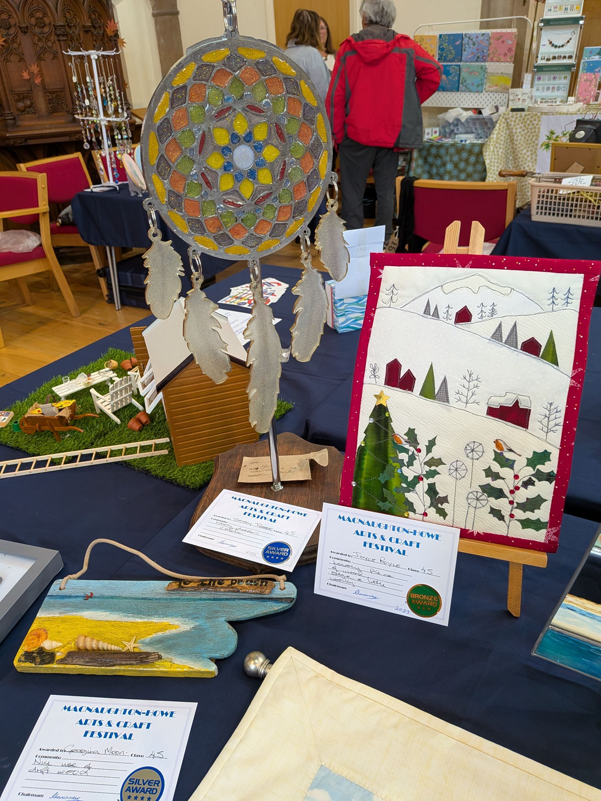 Craft fair table with handmade dream catcher, artwork, and signs on a blue tablecloth.