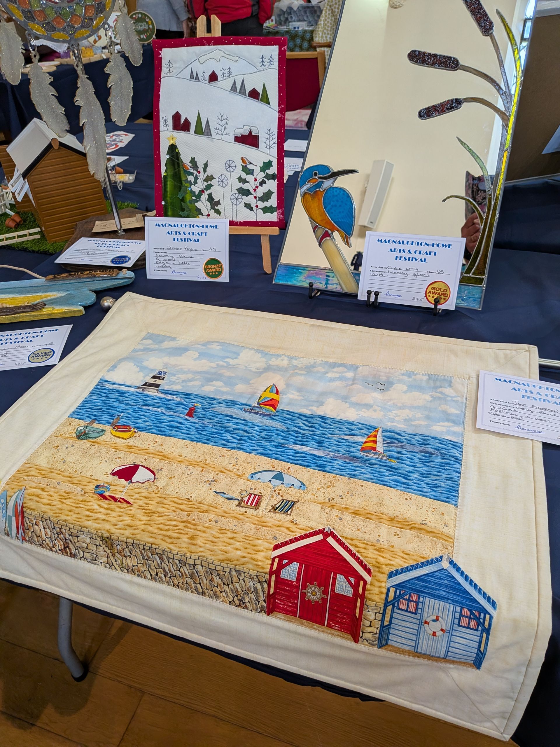 Quilted art display with a beach scene featuring colorful huts, sand, and ocean. Other artworks visible behind.