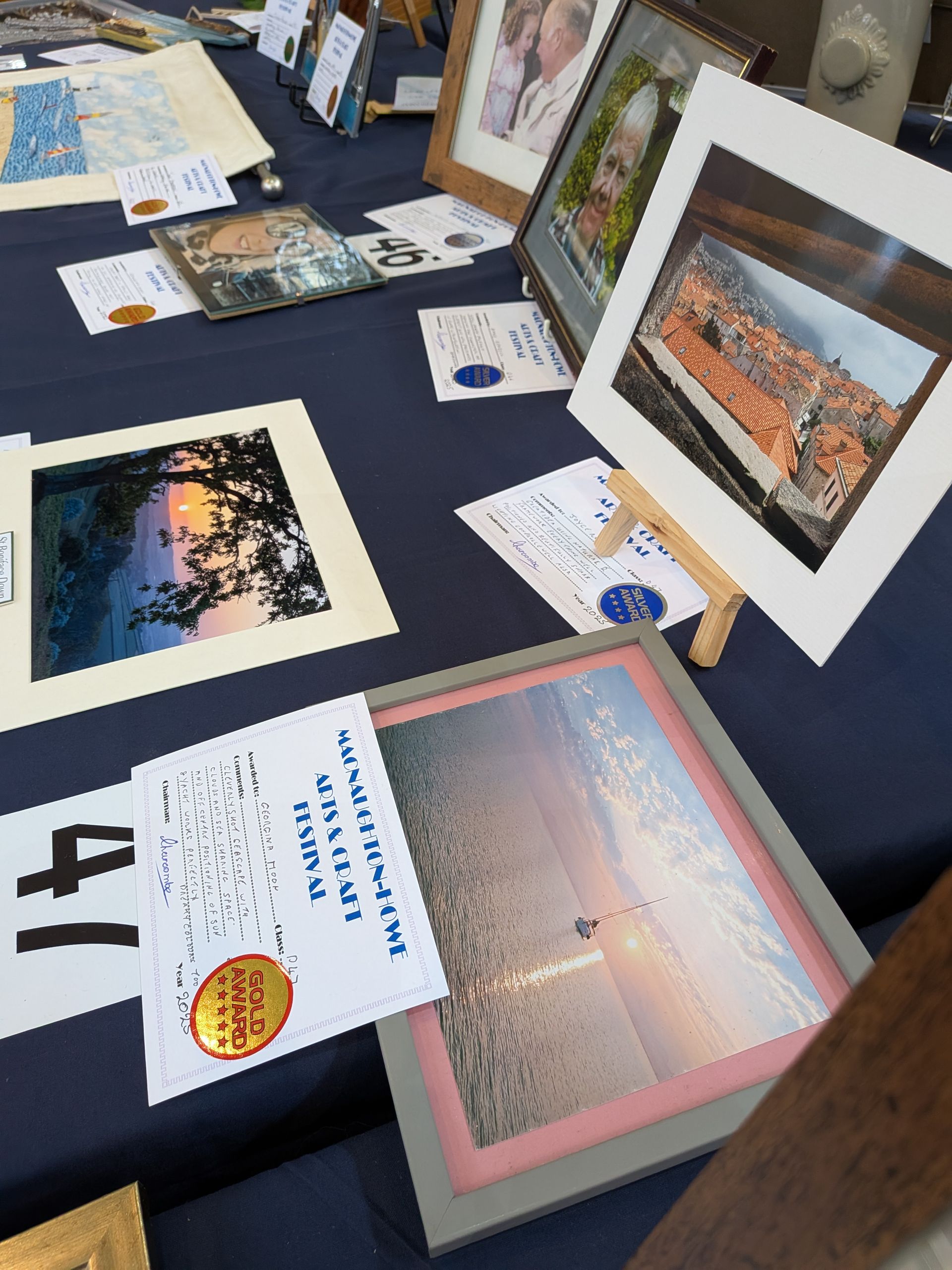 Artwork display on a blue tablecloth. Framed photos and prints with price tags and descriptive labels.