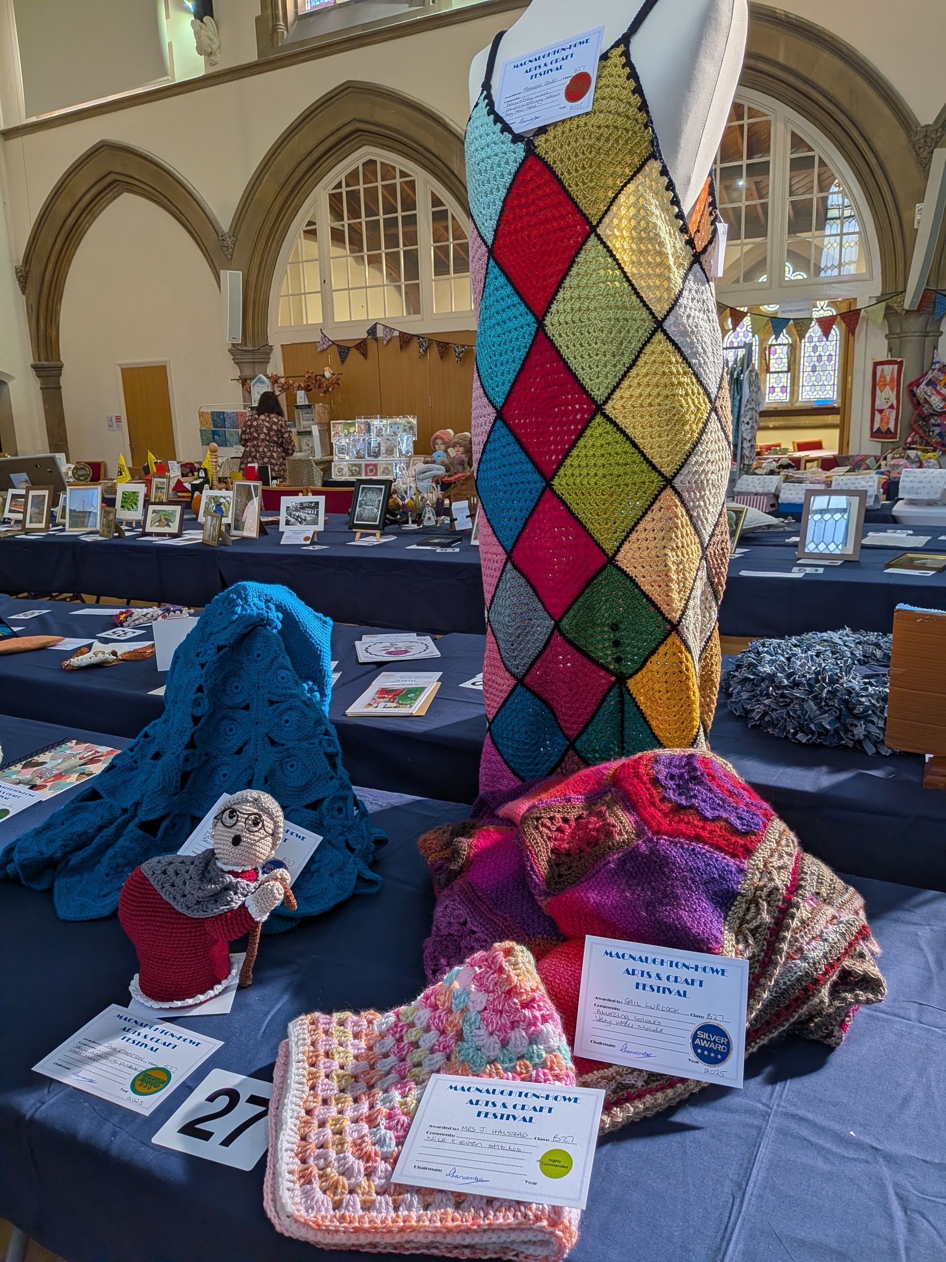 Display of handcrafted items, including a colorful, sequined dress, on a table in a bright room with arched windows.