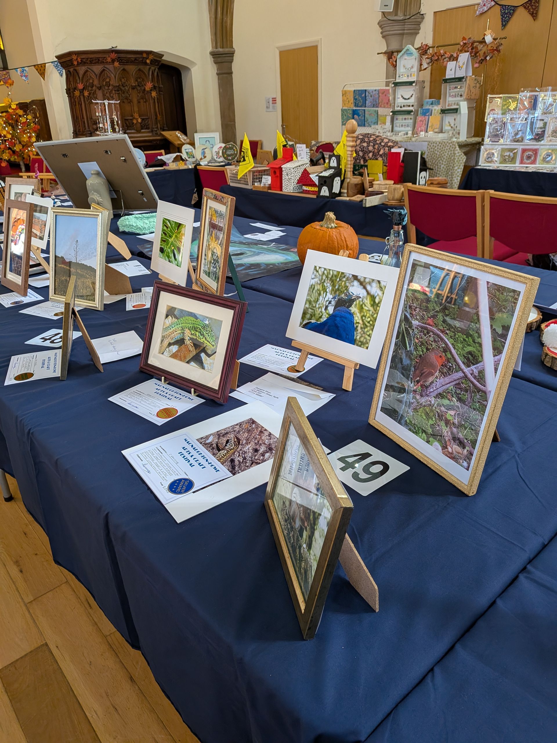 Art fair display: framed art prints on a blue tablecloth, with a pumpkin and other crafts in the background.