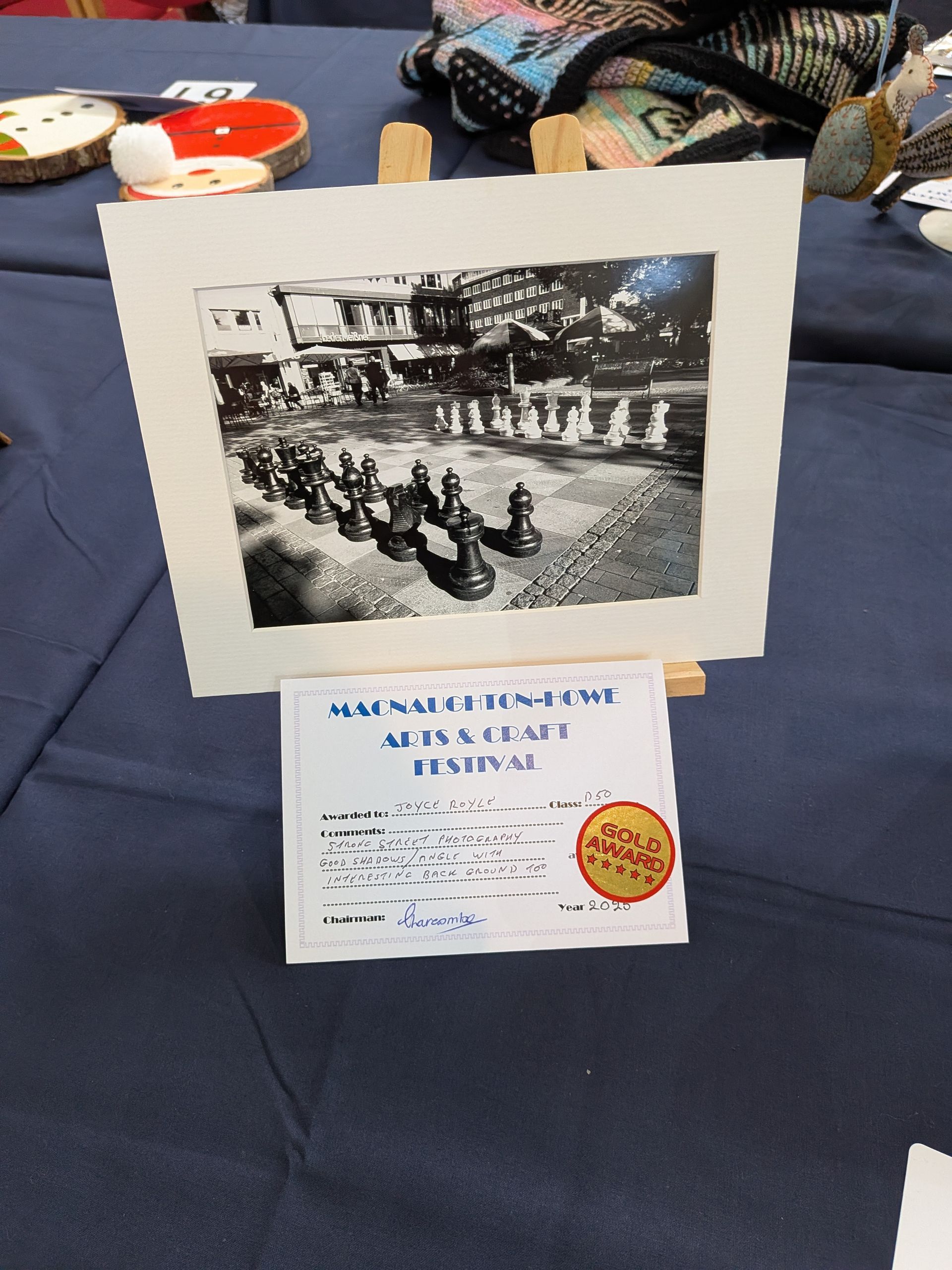 Black and white artwork on easel, depicting a row of chess pieces and architecture, with festival signage below.