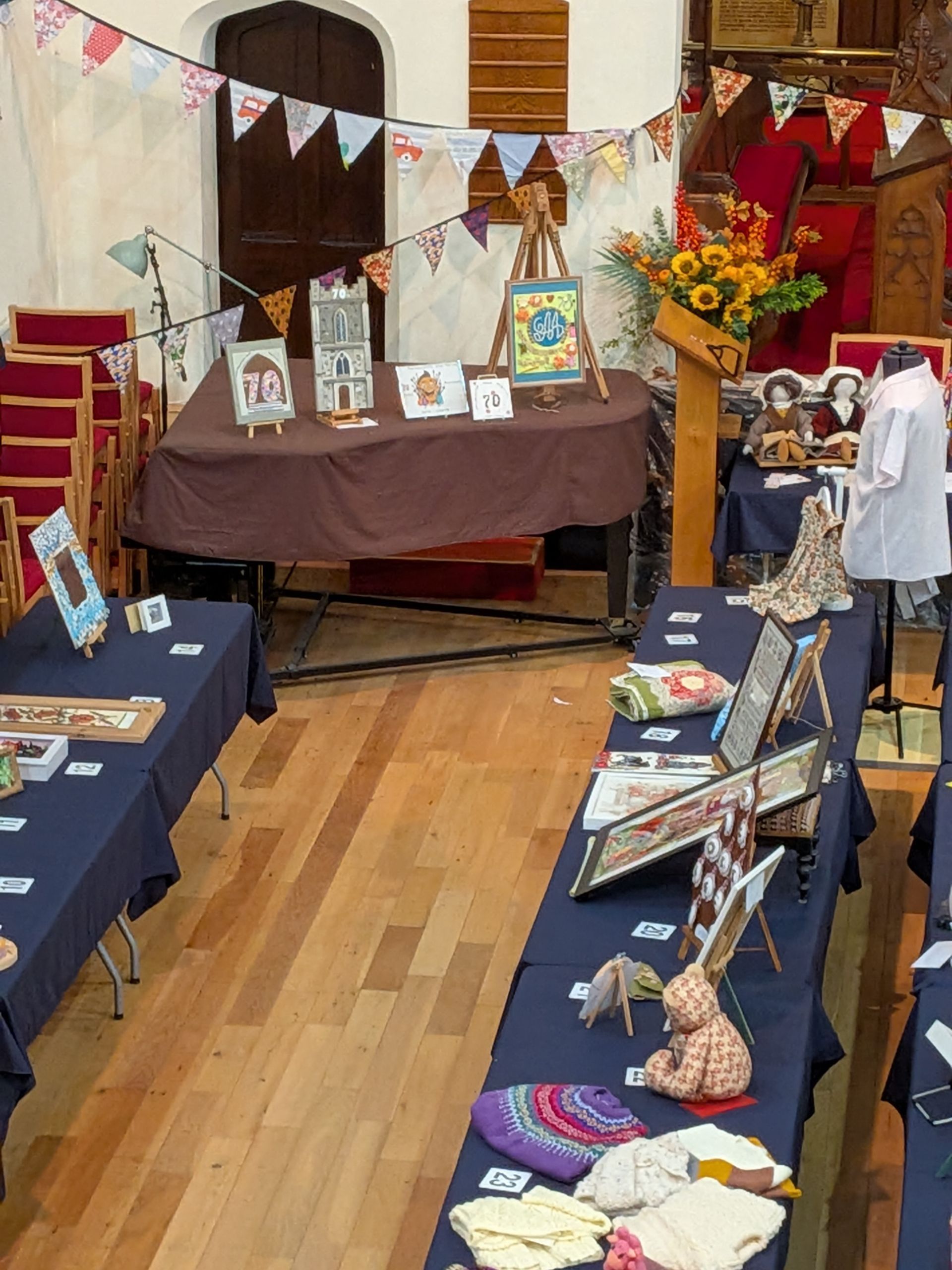 Craft fair inside a church: tables display artwork, crafts, and clothing; colorful bunting hangs overhead.