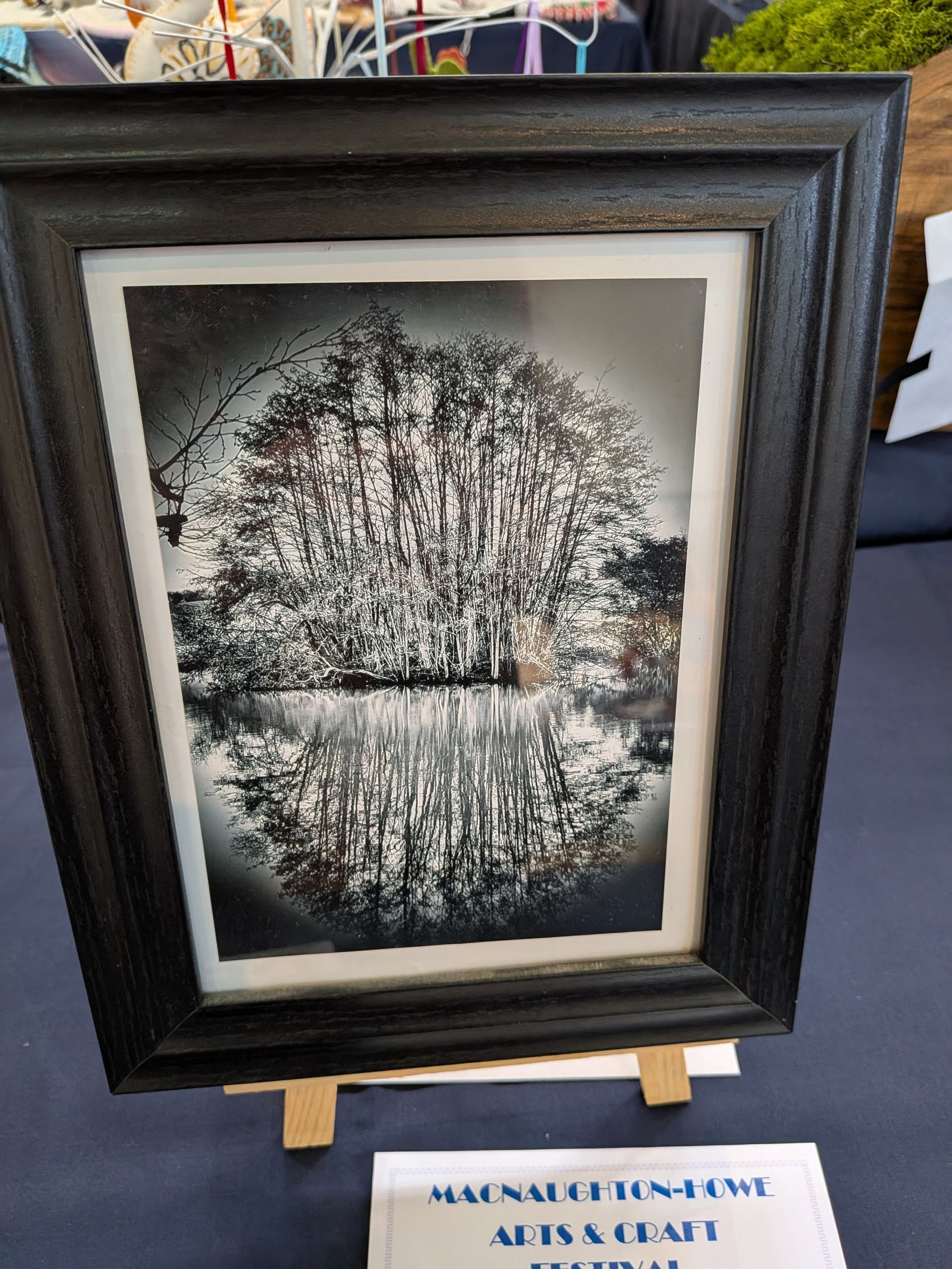 Black and white artwork of a tree reflected in water, framed and displayed on a stand.