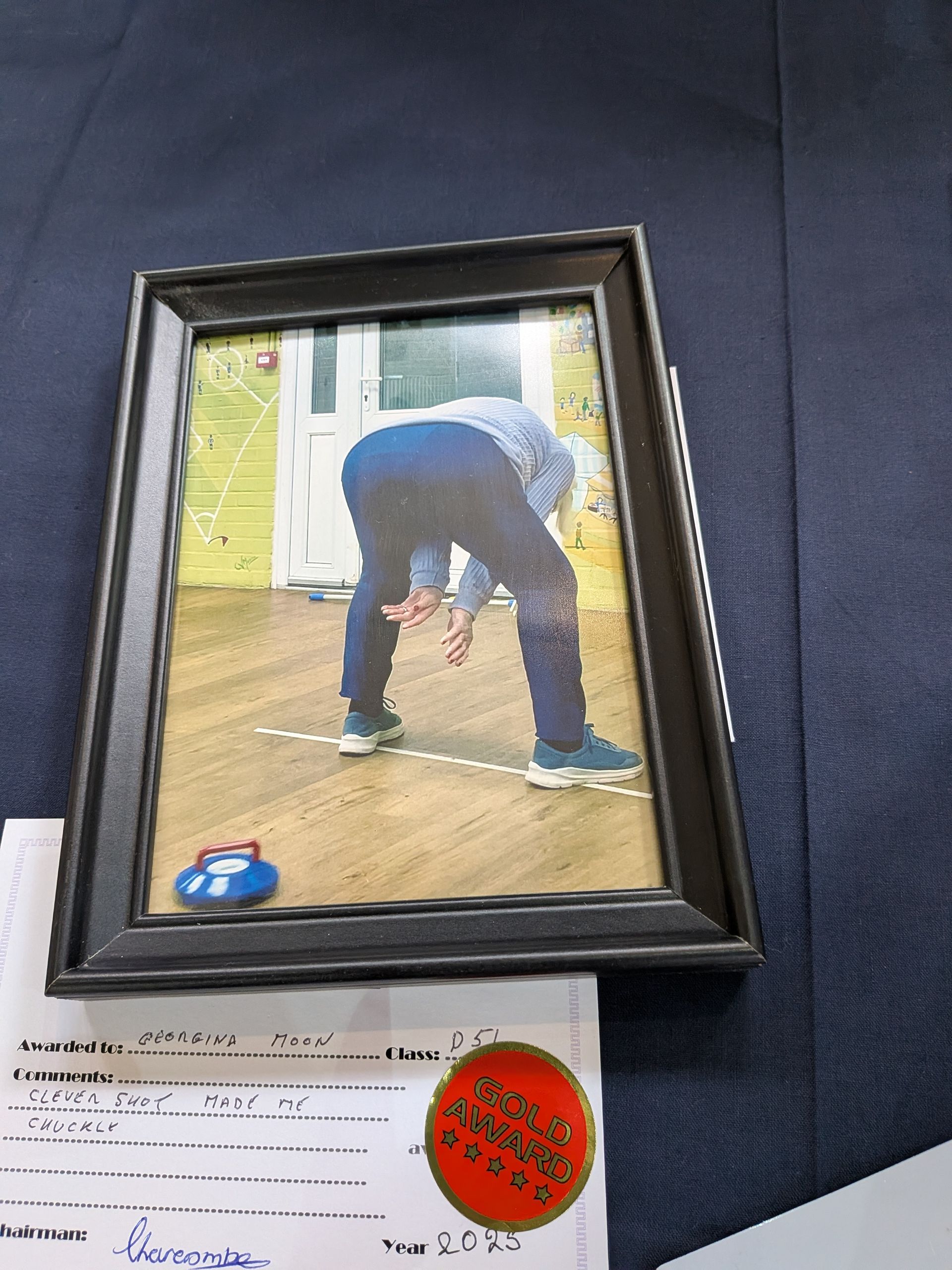 Person in blue jeans and shoes, bending over to sweep a curling stone on wooden porch. Framed photo.