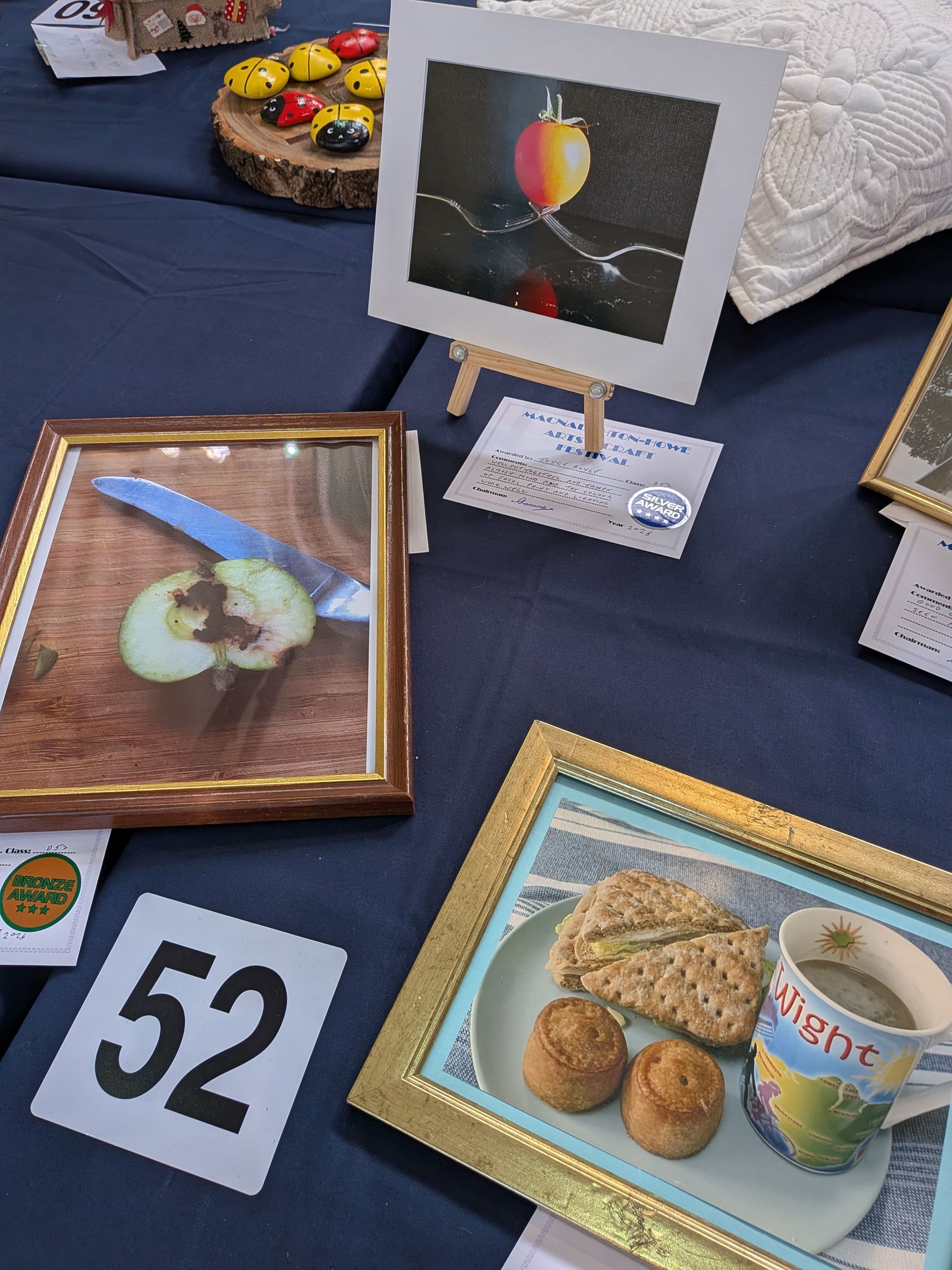 Artwork display: framed photos, painting on easel, food with number 52, on a dark blue surface.
