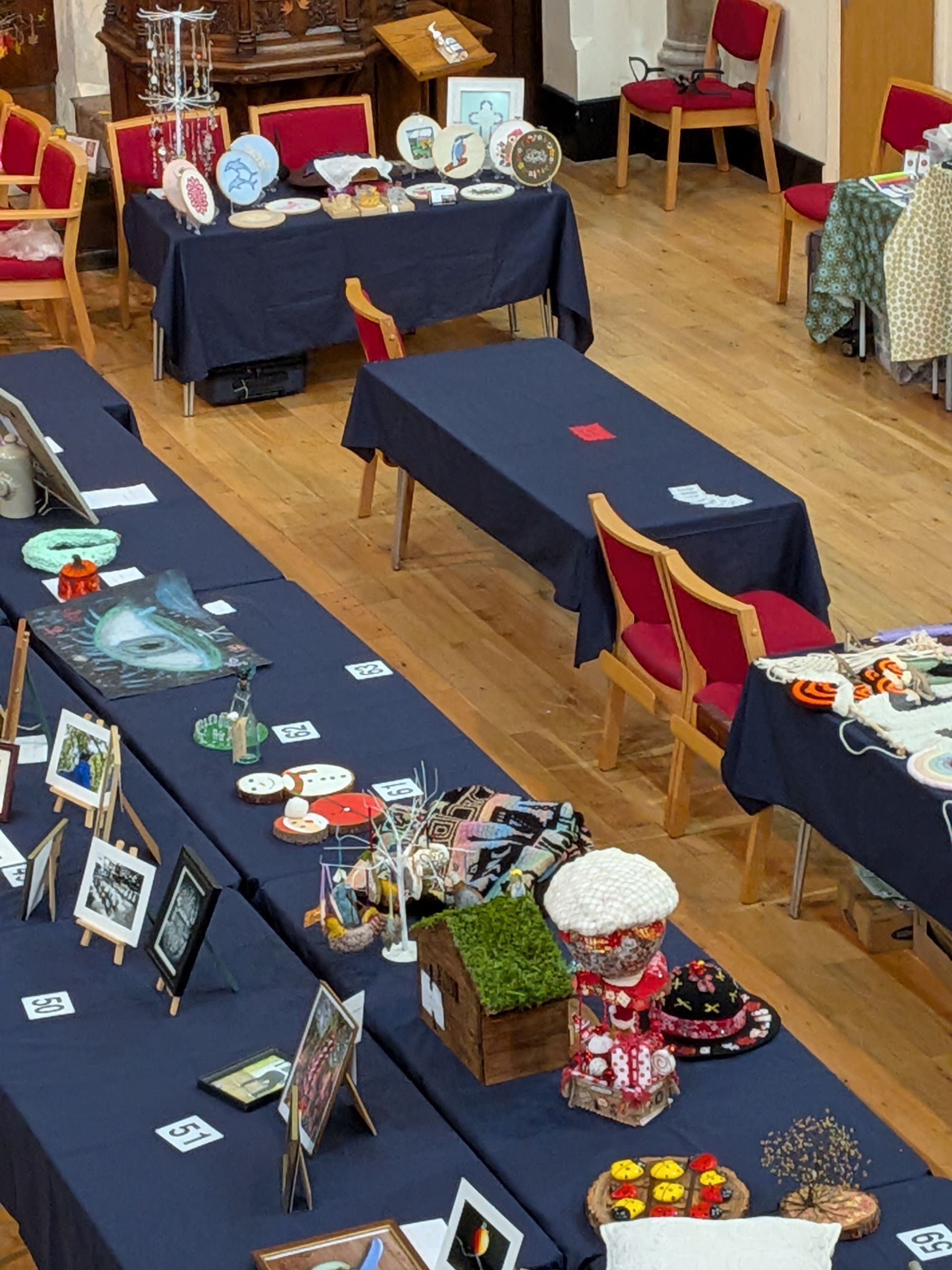 Craft fair tables with blue tablecloths, various crafts displayed. Wooden floor, red-backed chairs, and neutral lighting.