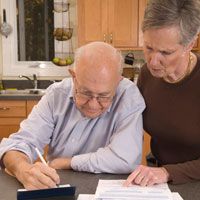 Elderly man with glasses writing as woman looks over his shoulder and points to a paper