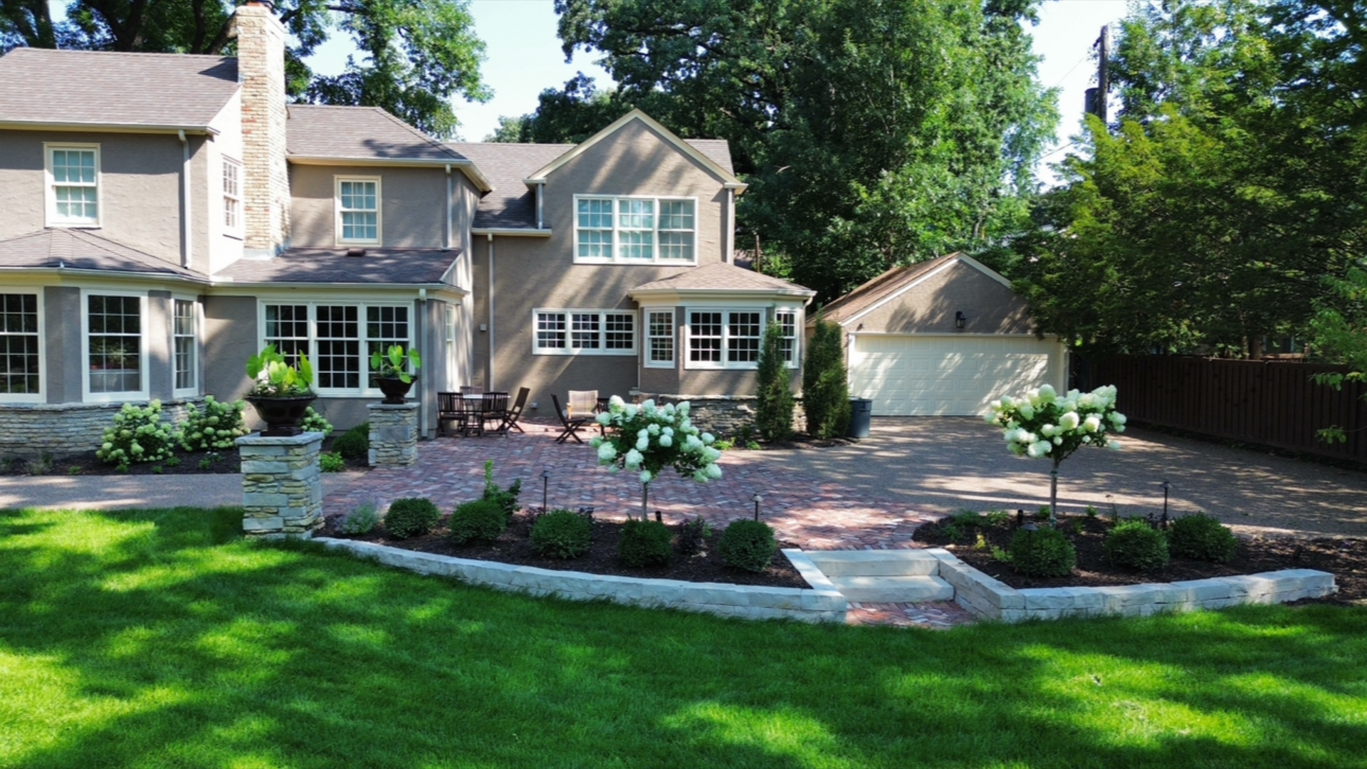 Two-story home with a brick driveway, manicured landscaping, and a detached garage on a sunny day.