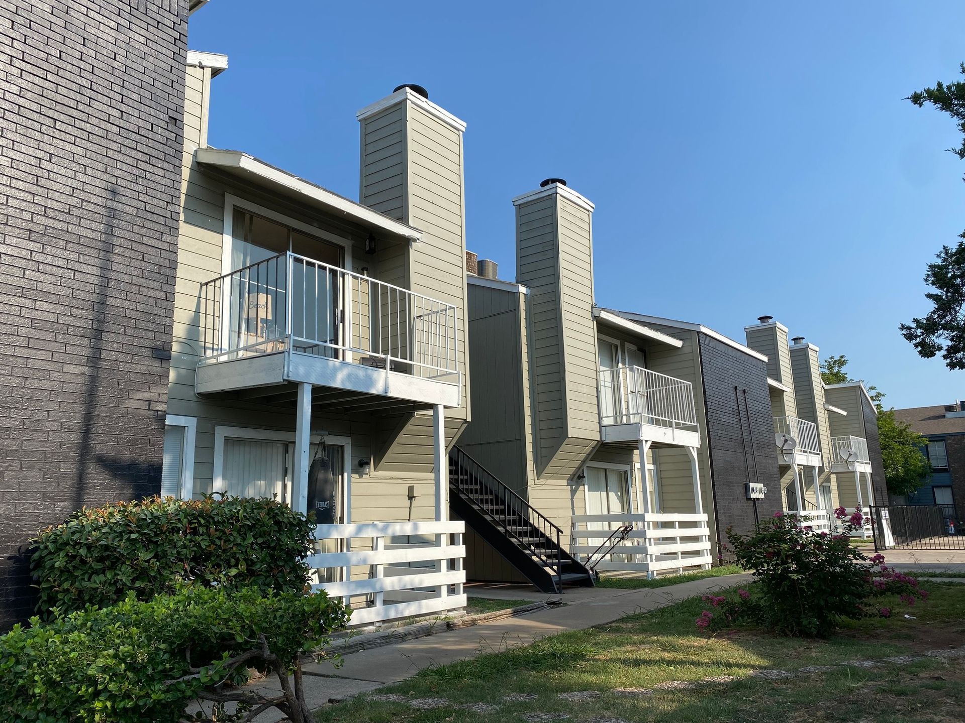 Apartment complex with gray siding, balconies, staircases, and brick exterior. Blue sky, sunny day.