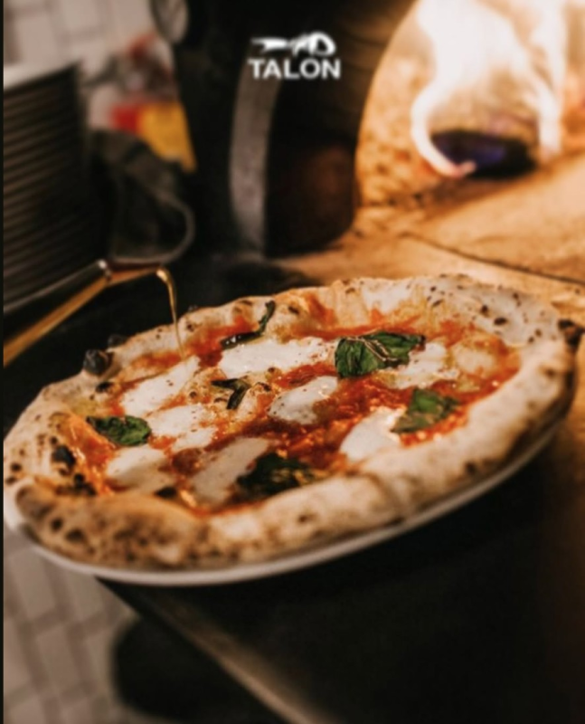 Una pizza Margherita cotta su un piatto davanti a un forno a legna; la crosta è dorata con basilico fresco e mozzarella.