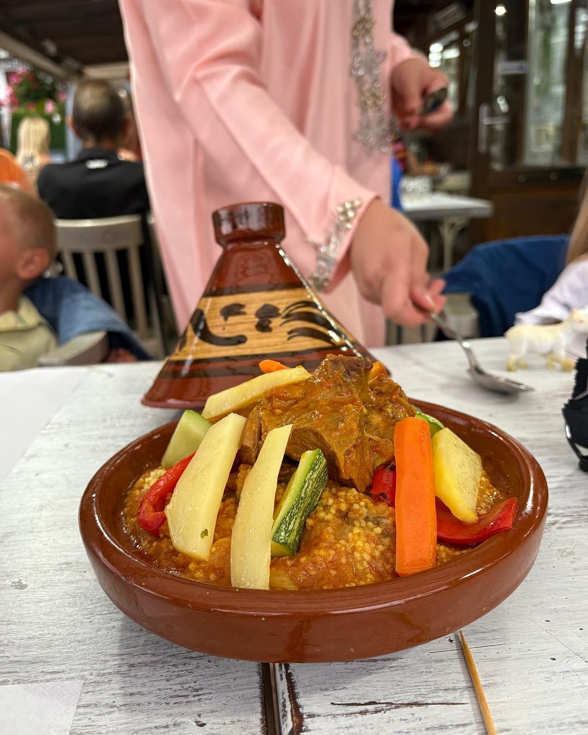 Tajine marocchino di agnello con verdure, servito su un piatto di terracotta e presentato da una persona in abito rosa.