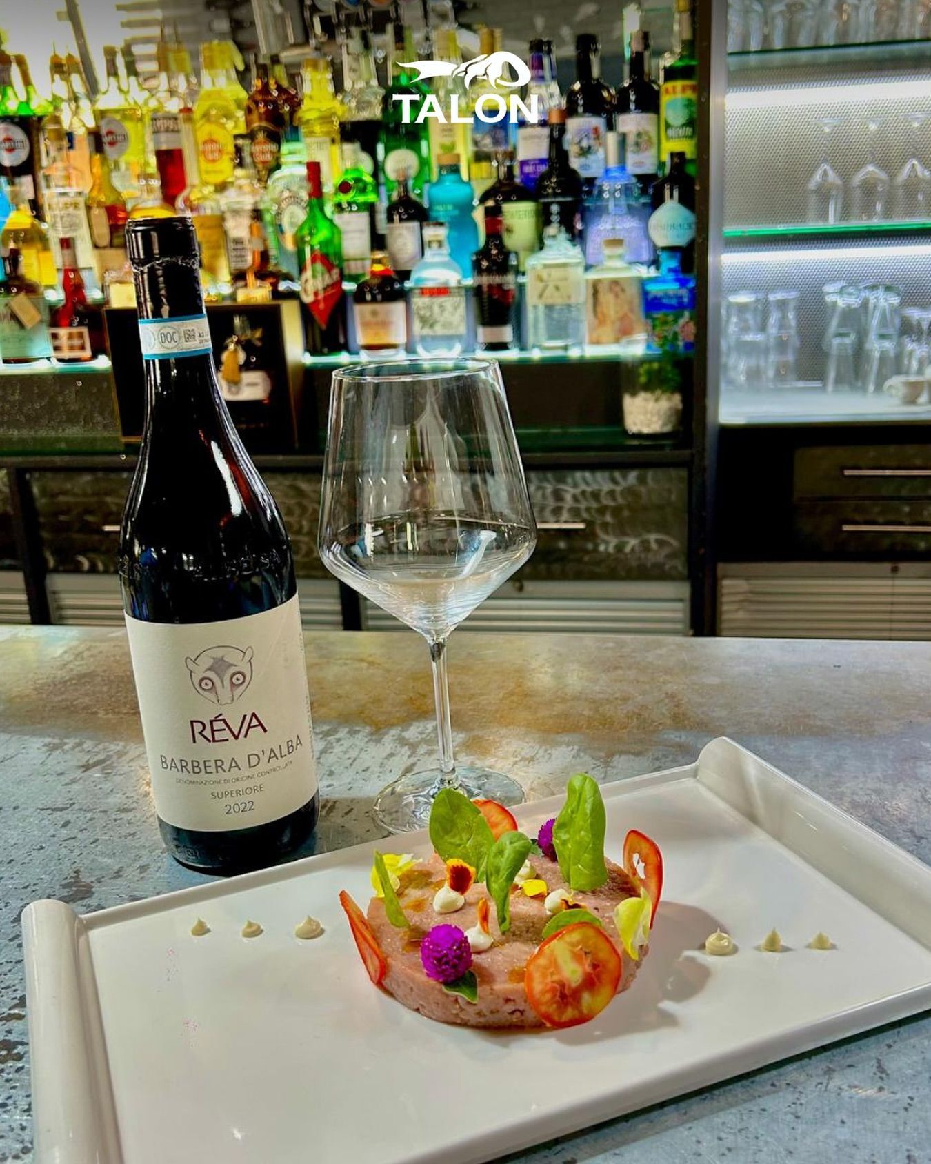 Una bottiglia di vino rosso, un bicchiere di vino e un piatto di gamberi su un bancone con bottiglie sullo sfondo.