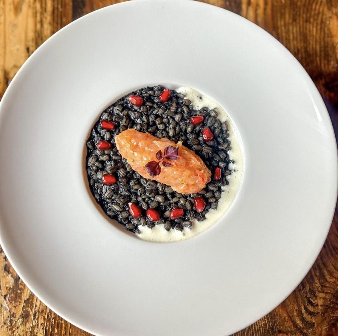 Risotto nero con salmone, semi di melograno e salsa cremosa su un piatto bianco.