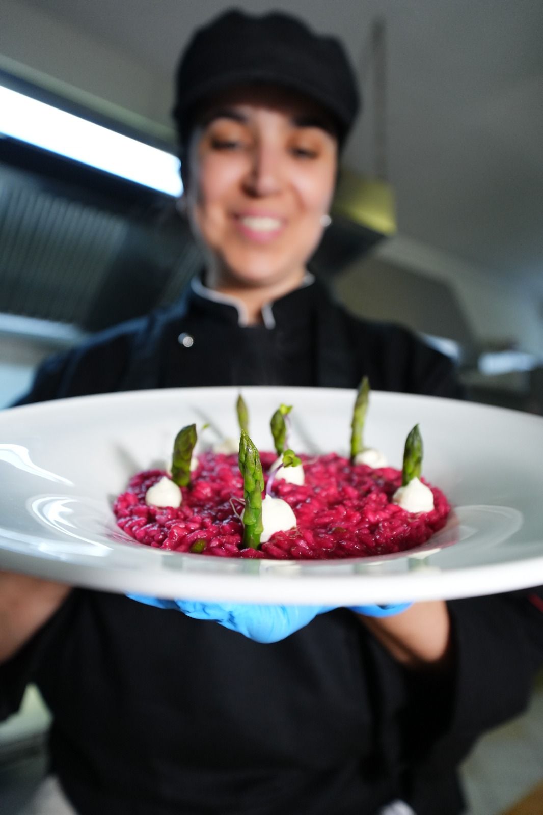 Uno chef sorridente in cucina presenta un piatto di risotto rosa con asparagi.