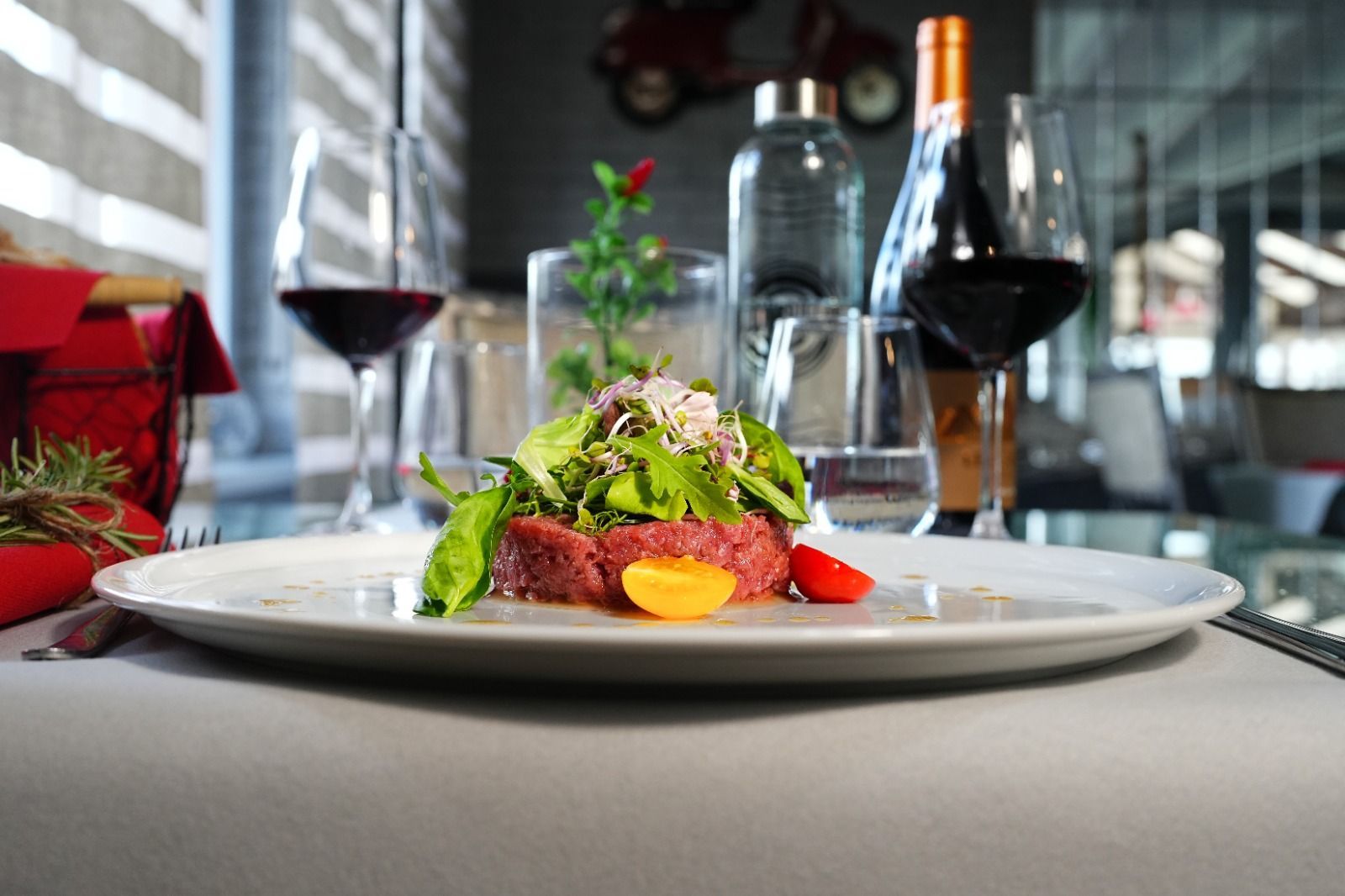 Piatto di tartare di manzo con insalata, pomodori, bicchieri di vino rosso, bottiglia d'acqua e ambiente da ristorante.