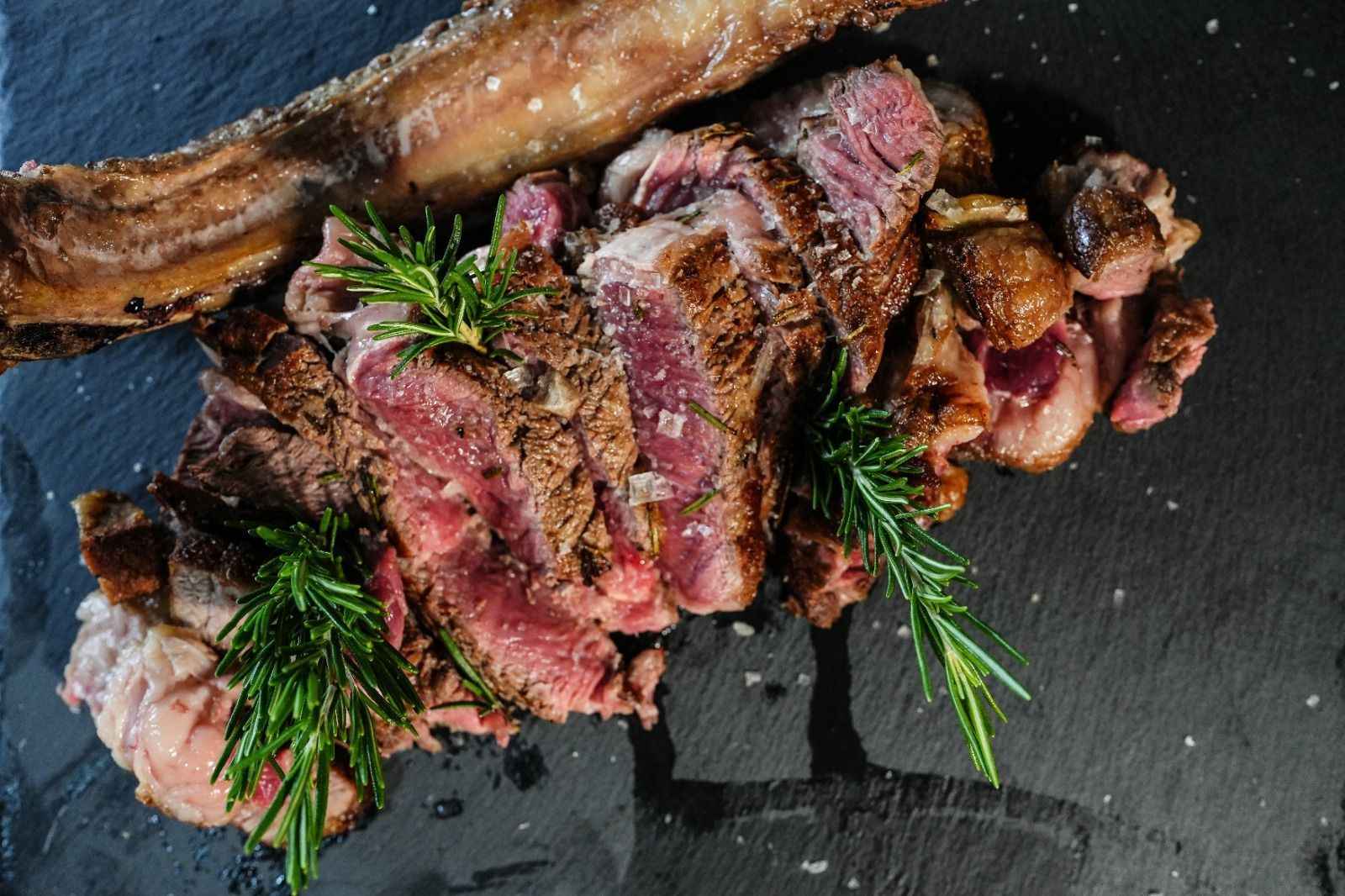 Bistecca di manzo tagliata a fette, con l'osso, guarnita con rametti di rosmarino e sale, su una superficie di ardesia scura.