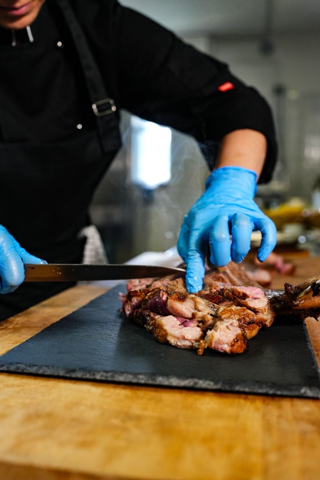 Cuoco in uniforme nera che affetta carne arrostita su una tavola di ardesia, indossando guanti blu.