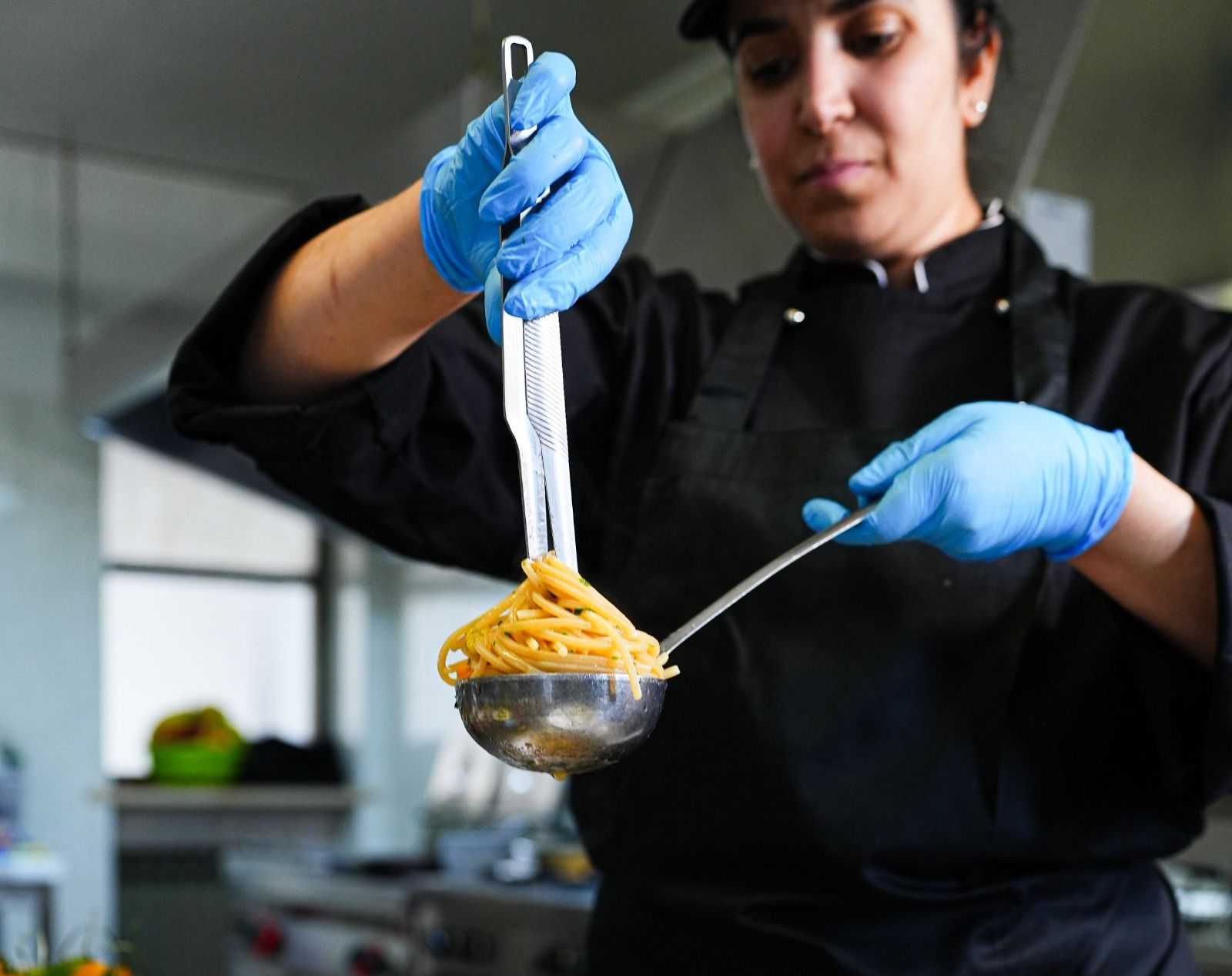 Cuoco in uniforme nera e guanti blu che serve la pasta con un mestolo in cucina.
