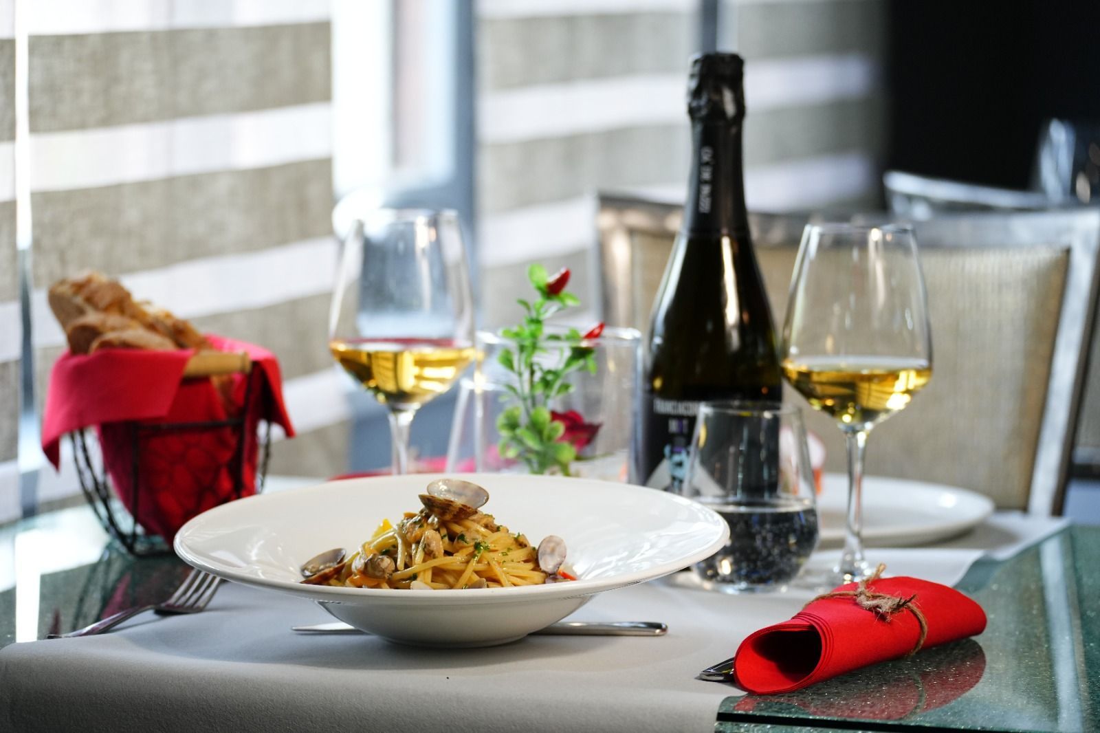 Piatto di pasta con bicchieri di vino, una bottiglia e pane in un ambiente da ristorante.