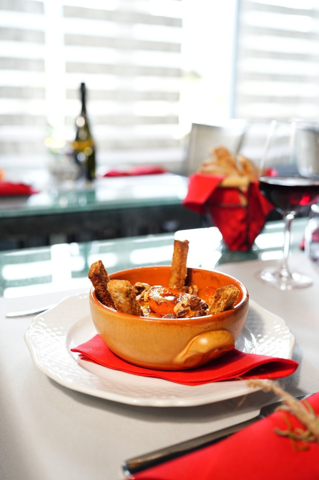 Ciotola di cibo con grissini su un piatto bianco, tovagliolo rosso, bicchiere di vino e finestra sullo sfondo.