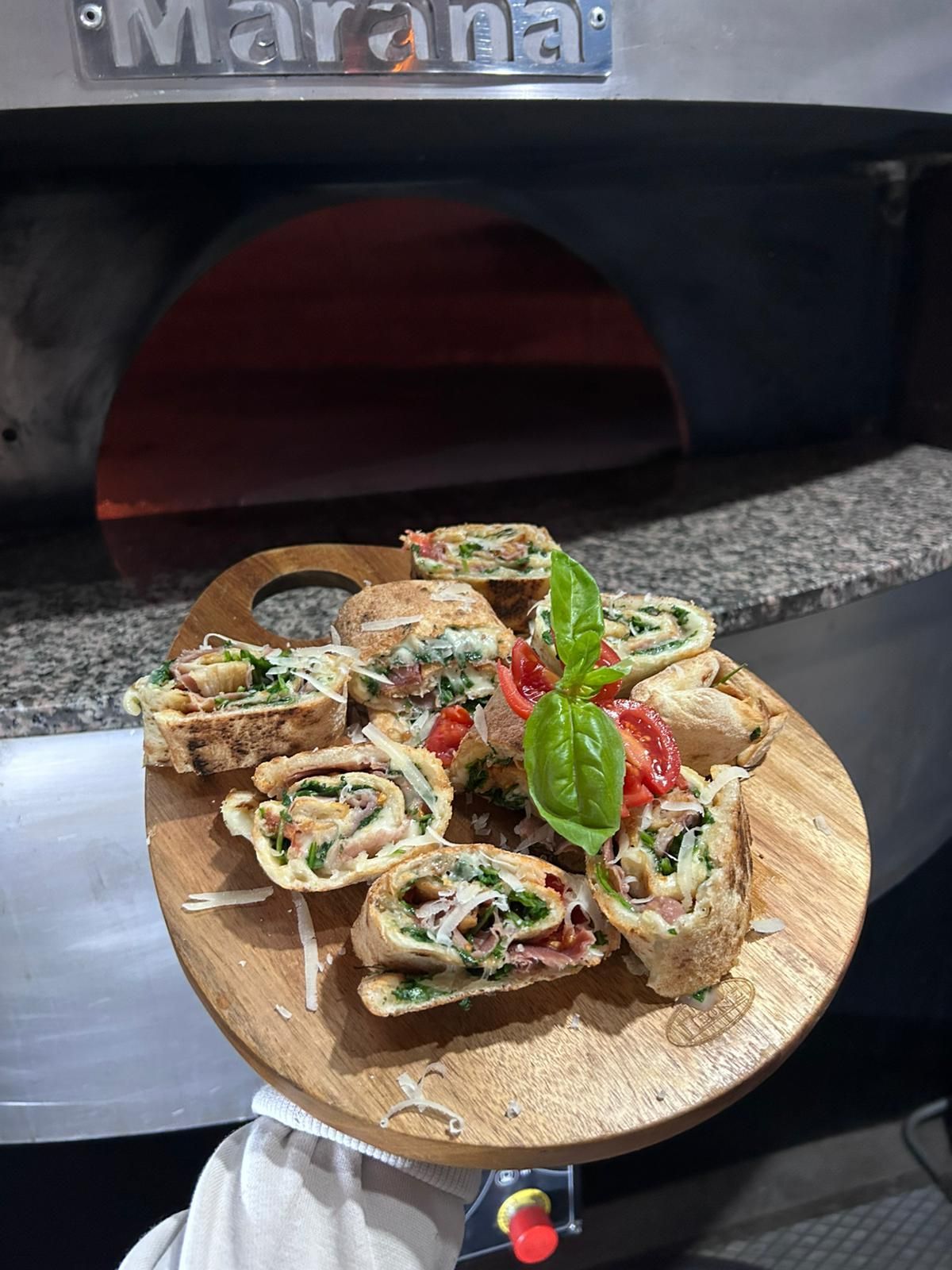 Tagliere di legno con piadine tagliate a fette, guarnite con pomodori e basilico, davanti a un forno per pizza.