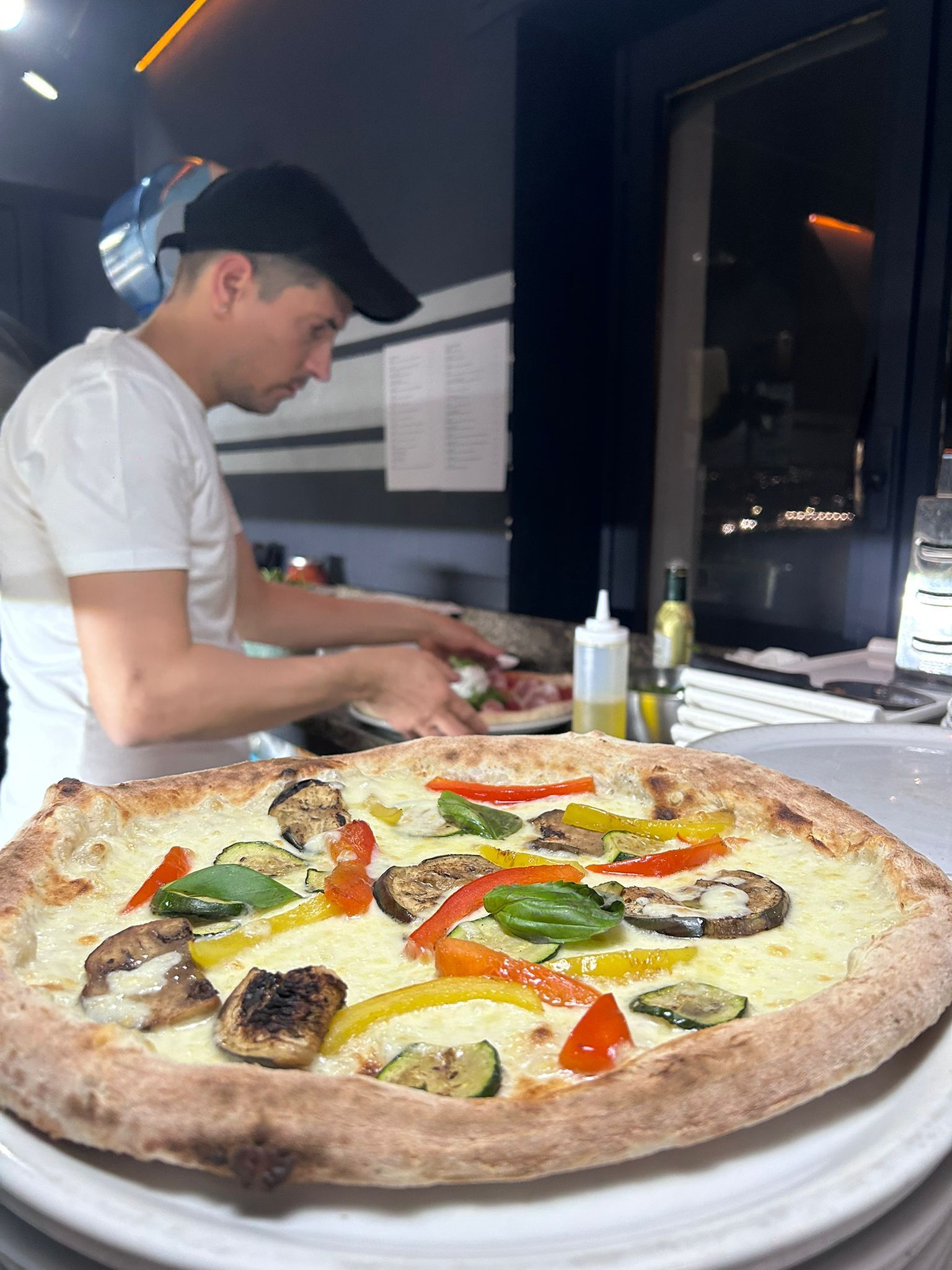 Un pizzaiolo prepara i condimenti su una pizza con la superficie bianca, con le verdure colorate in vista, in una cucina.