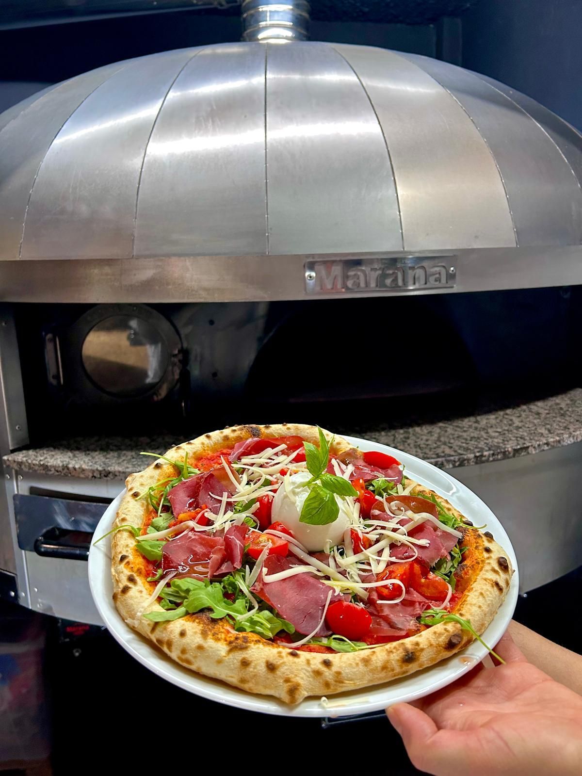 Pizza su un piatto, tenuta in alto davanti a un forno per pizza. La pizza è condita ed è pronta da mangiare.